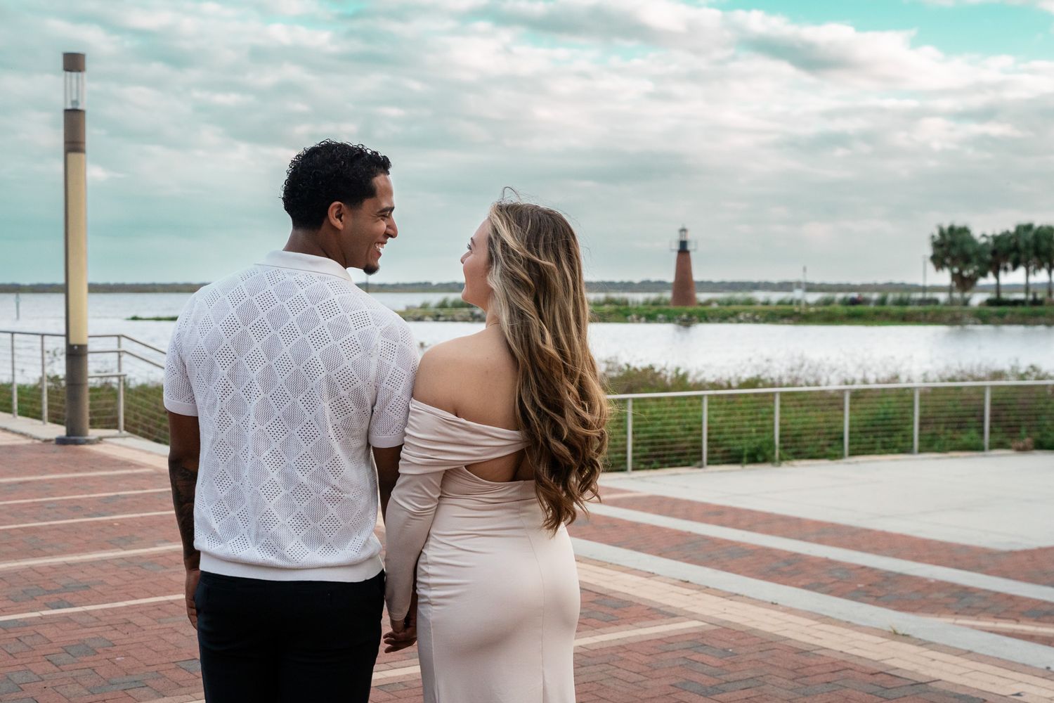 Couple holding hands walking along waterfront promenade with lighthouse in background.