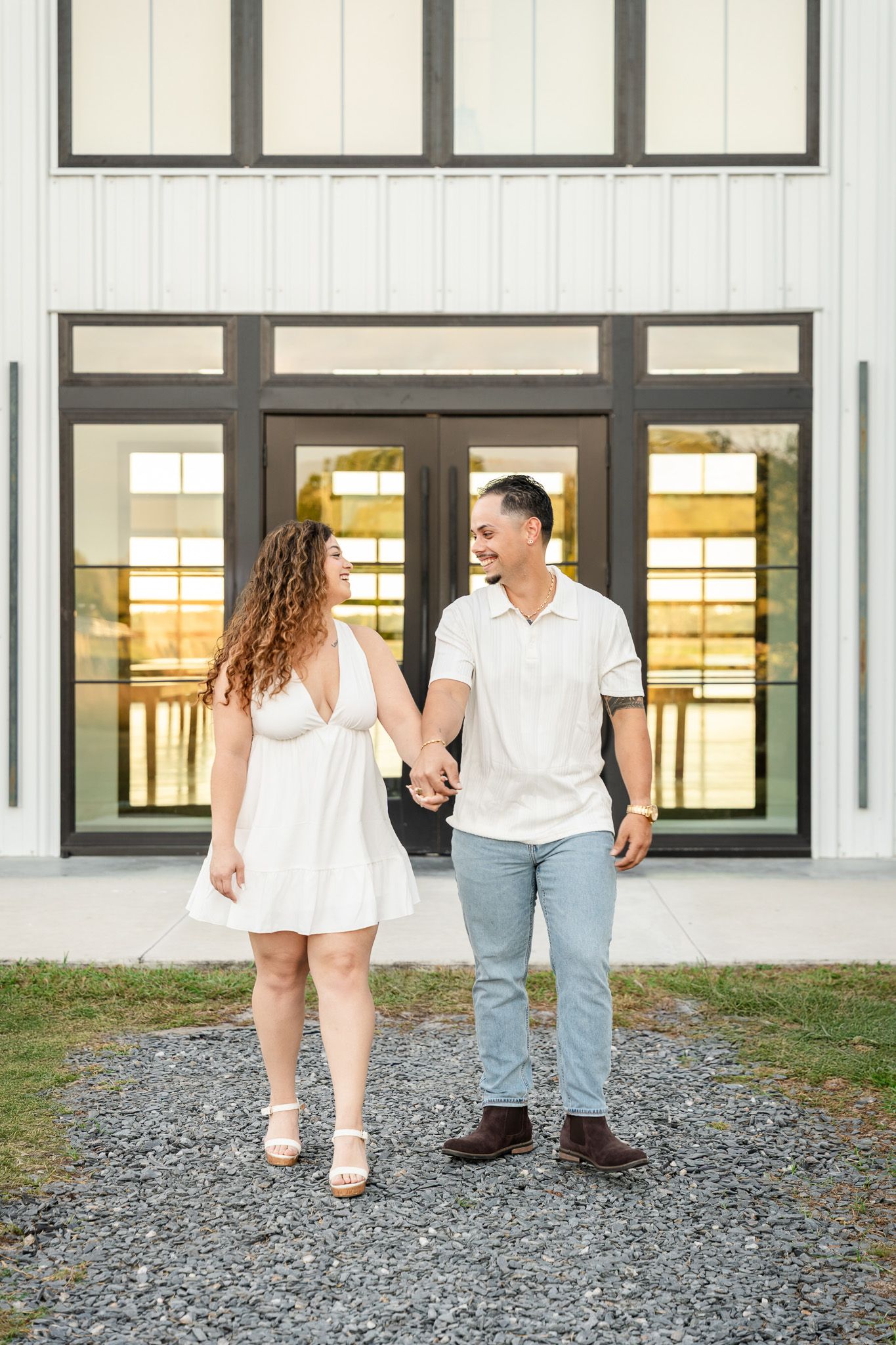 Happy couple holds hands walking in front of modern white barn building.
