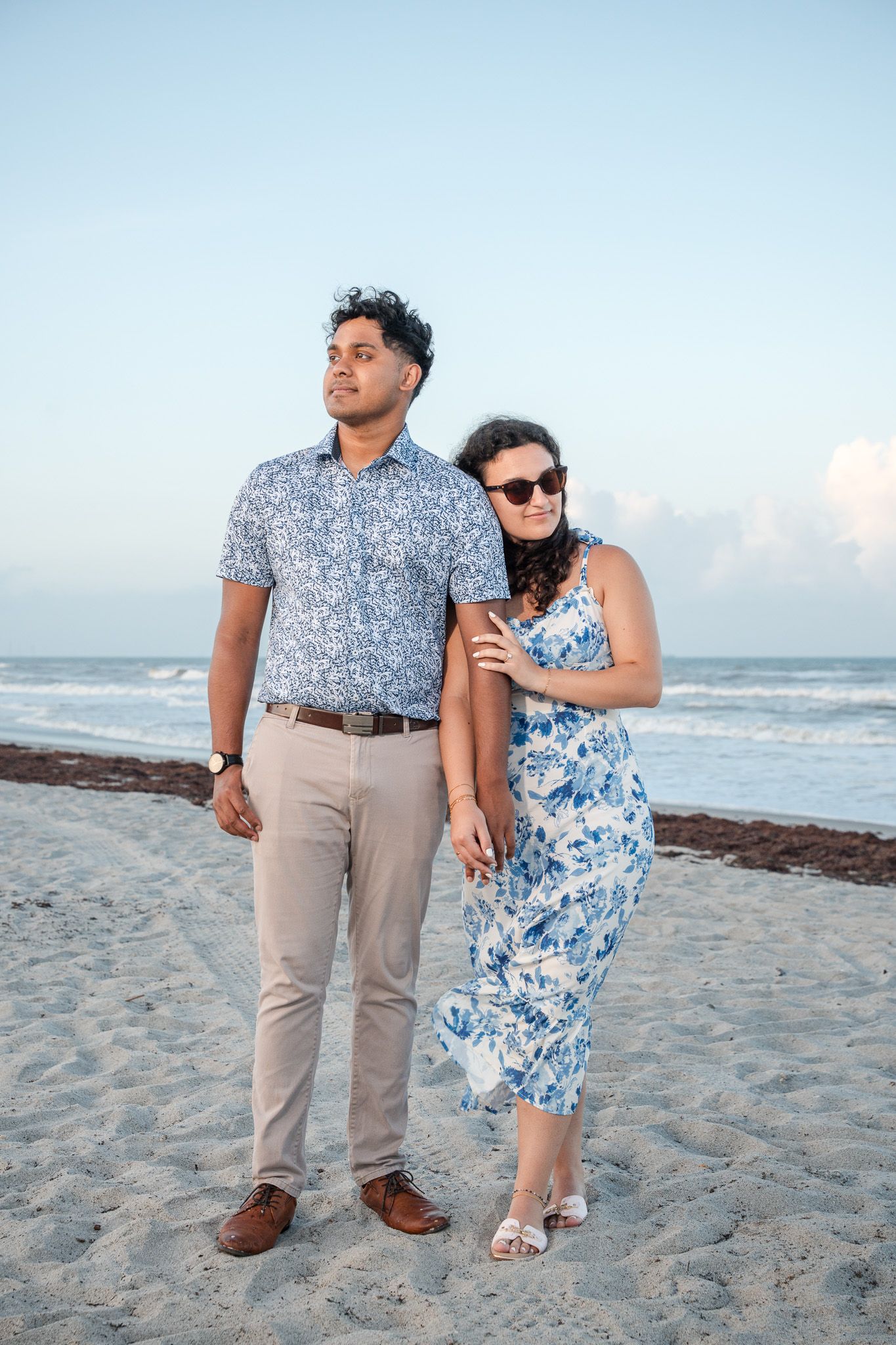 Couple posing on sandy beach at dusk, man in floral shirt and khakis, woman in blue floral dress and sunglasses.