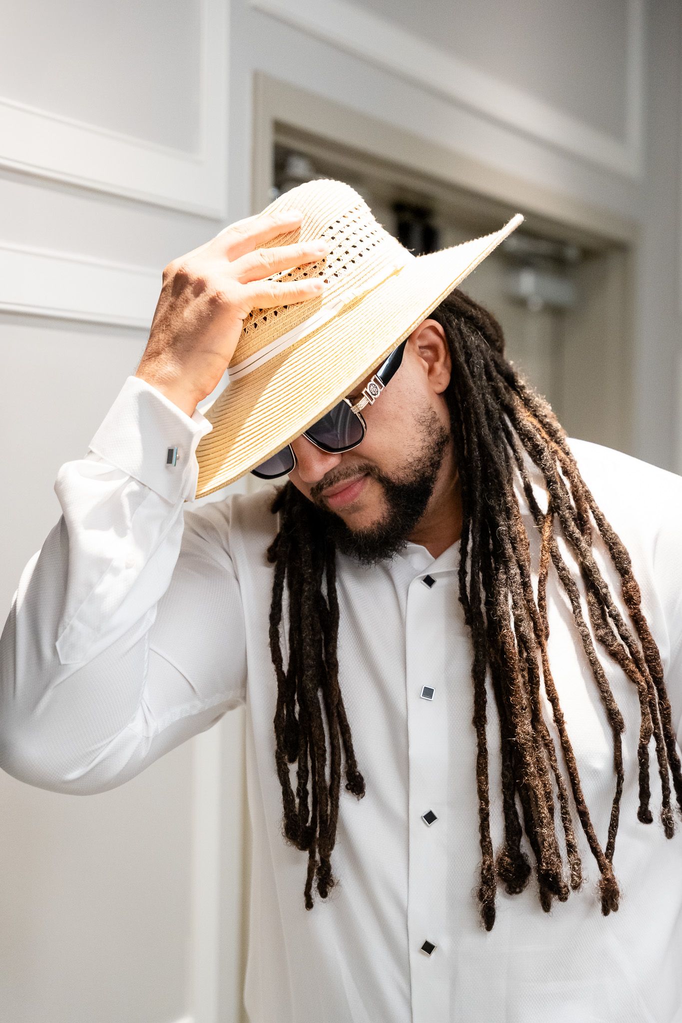 Man with long dreadlocks adjusts straw hat while wearing white shirt and sunglasses indoors.