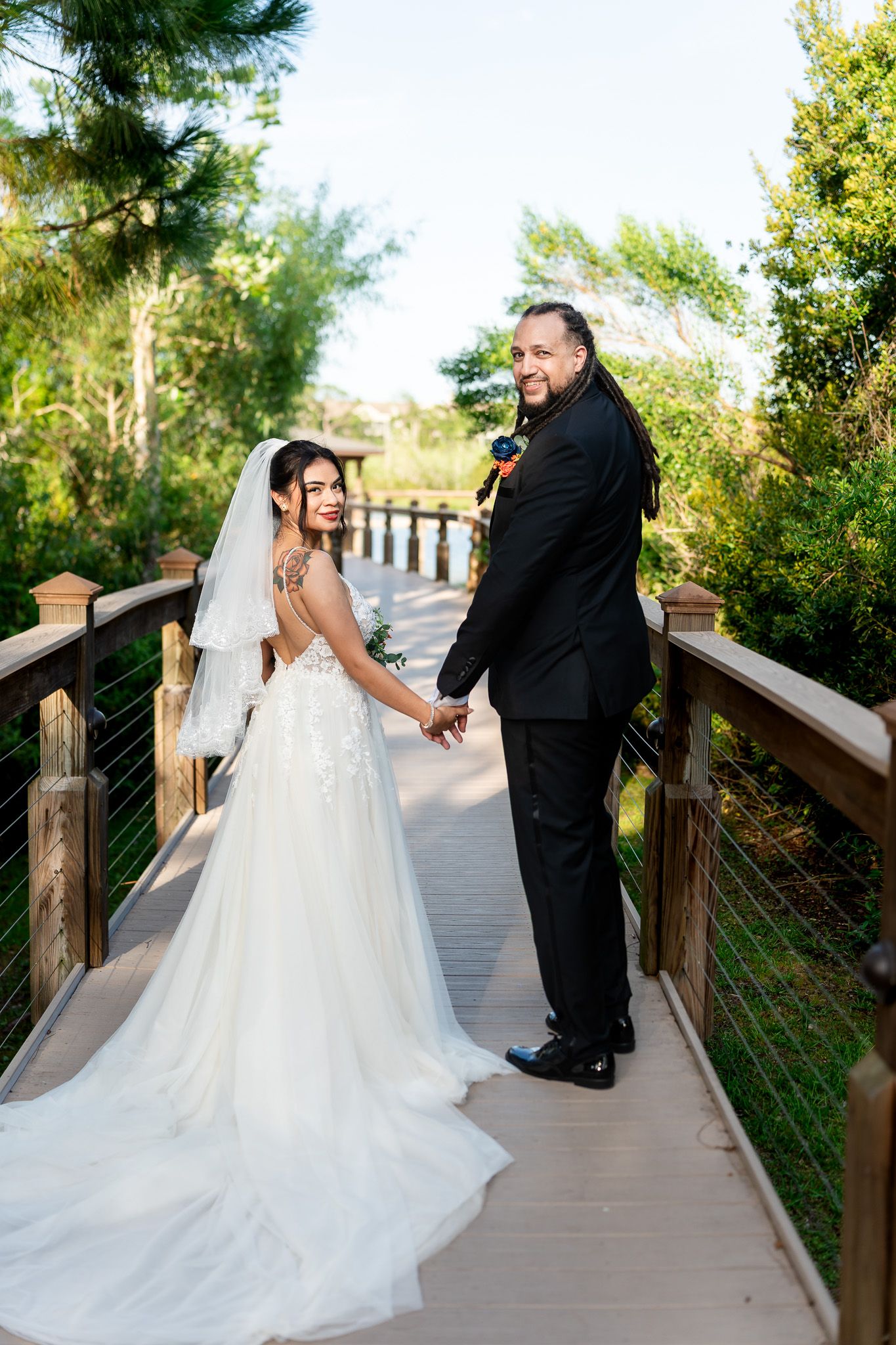 Bride and groom holding hands on wooden bridge surrounded by lush greenery.