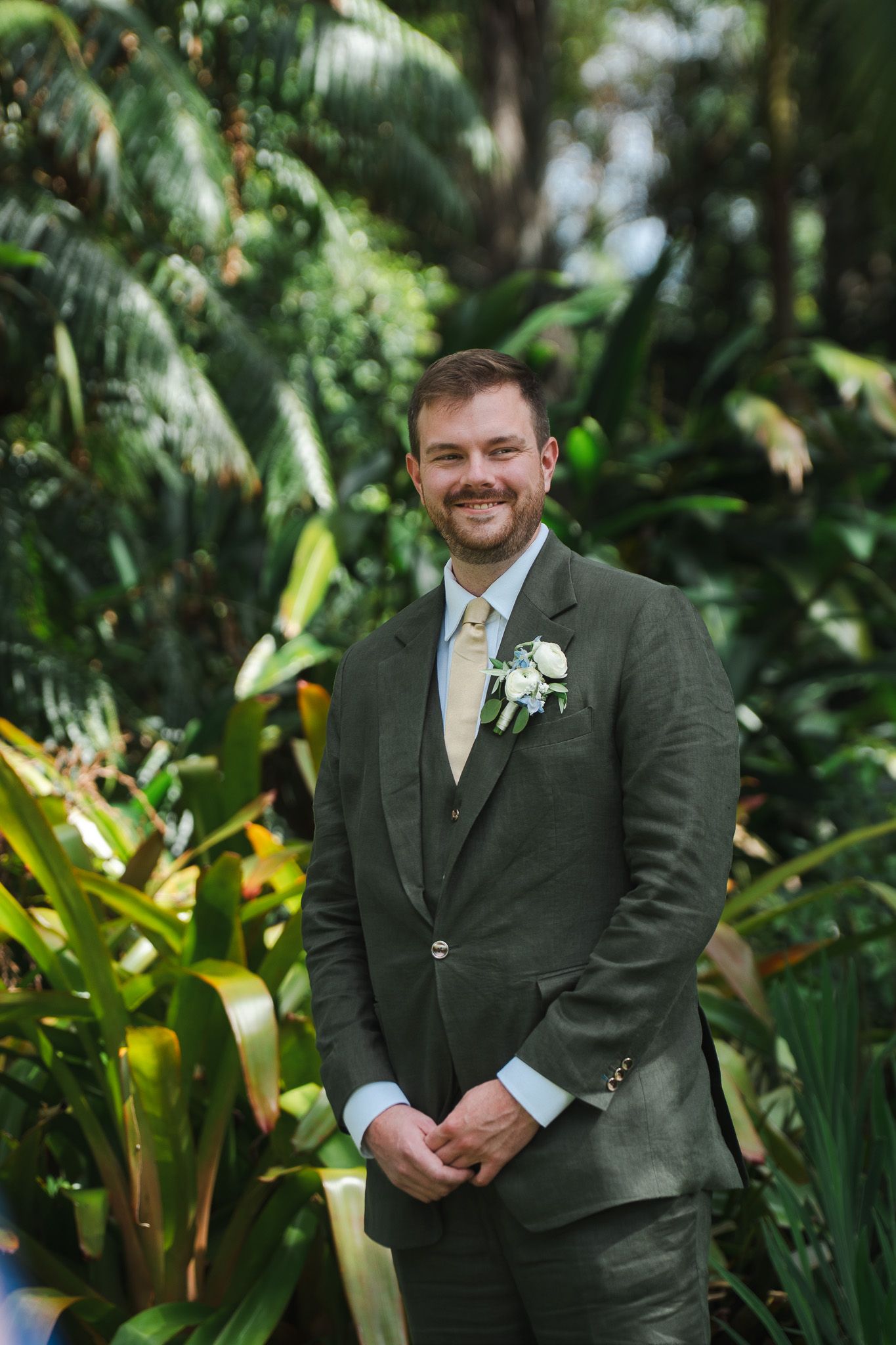 Handsome groom in olive green suit with boutonniere smiles before outdoor garden wedding ceremony.