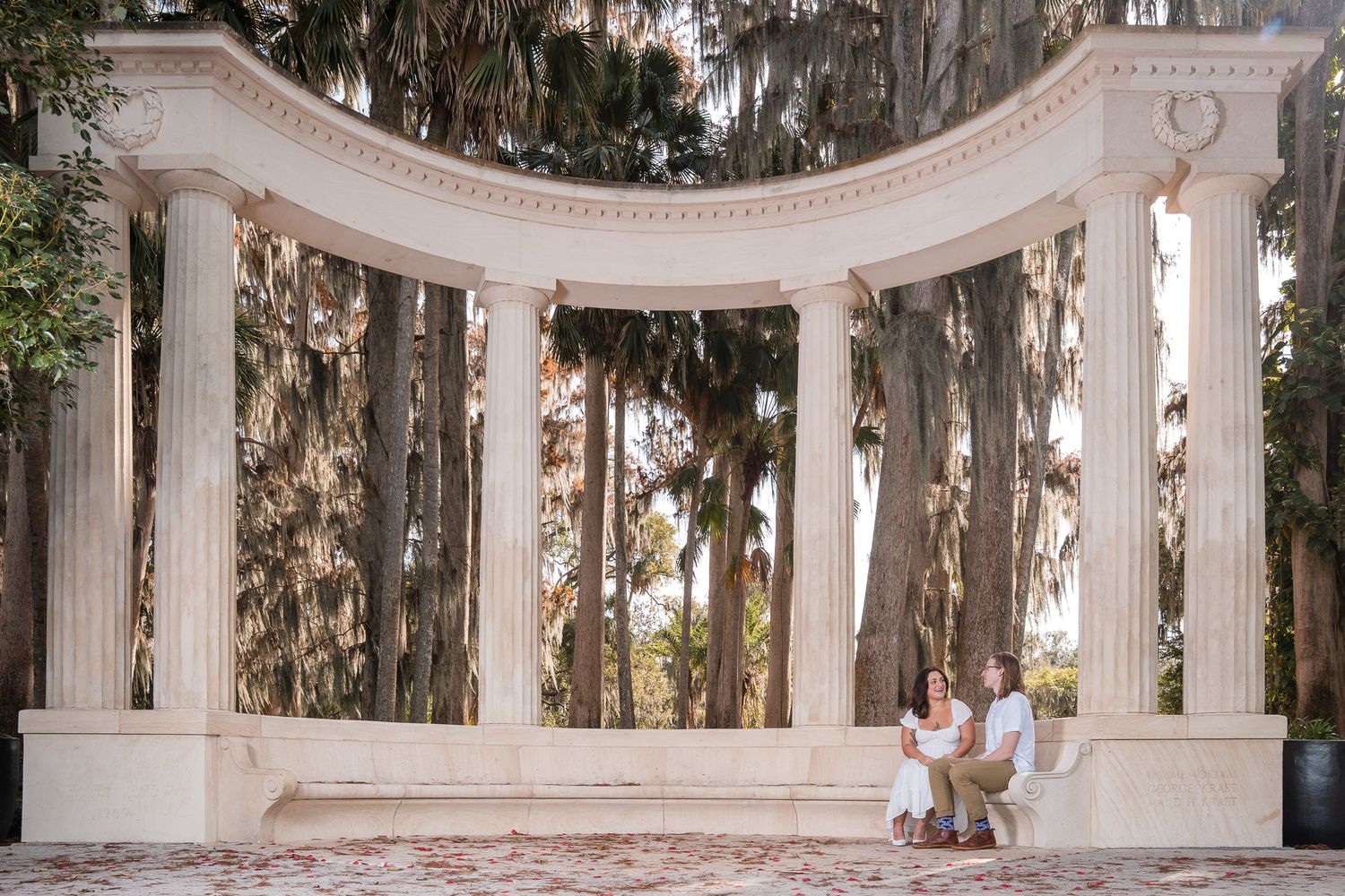 Couple sits closely together under curved white stone colonnade surrounded by lush Spanish moss-draped trees.