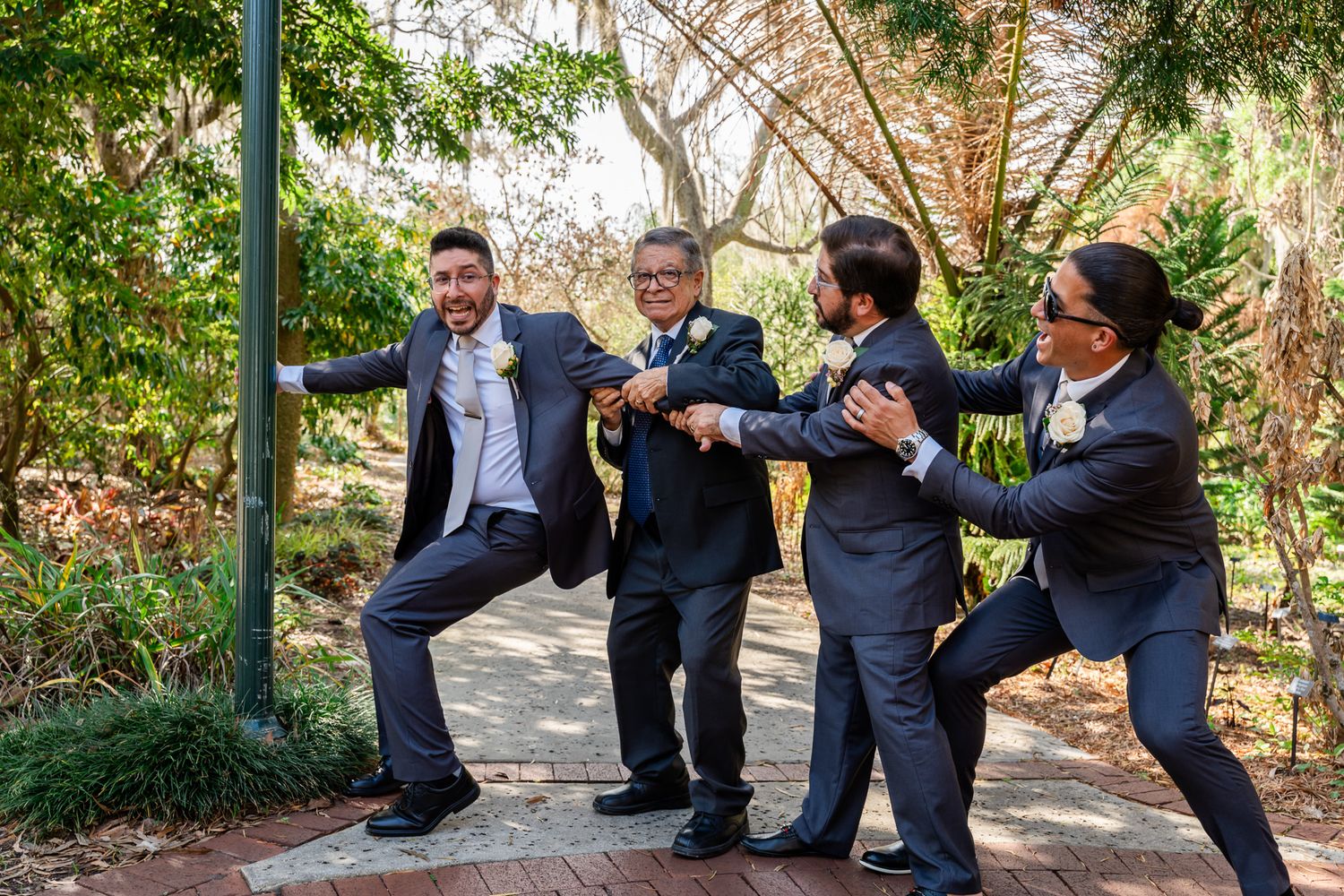 Groomsmen in gray suits laugh and strike fun poses together along an outdoor garden pathway.