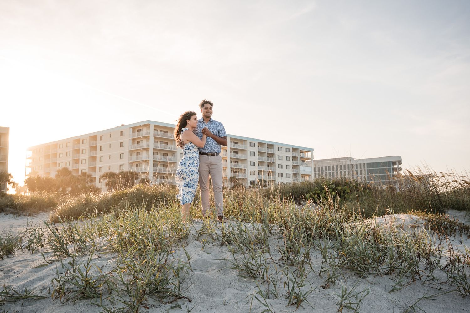 Happy couple stands together on a sandy dune with coastal buildings and sunset sky behind them.