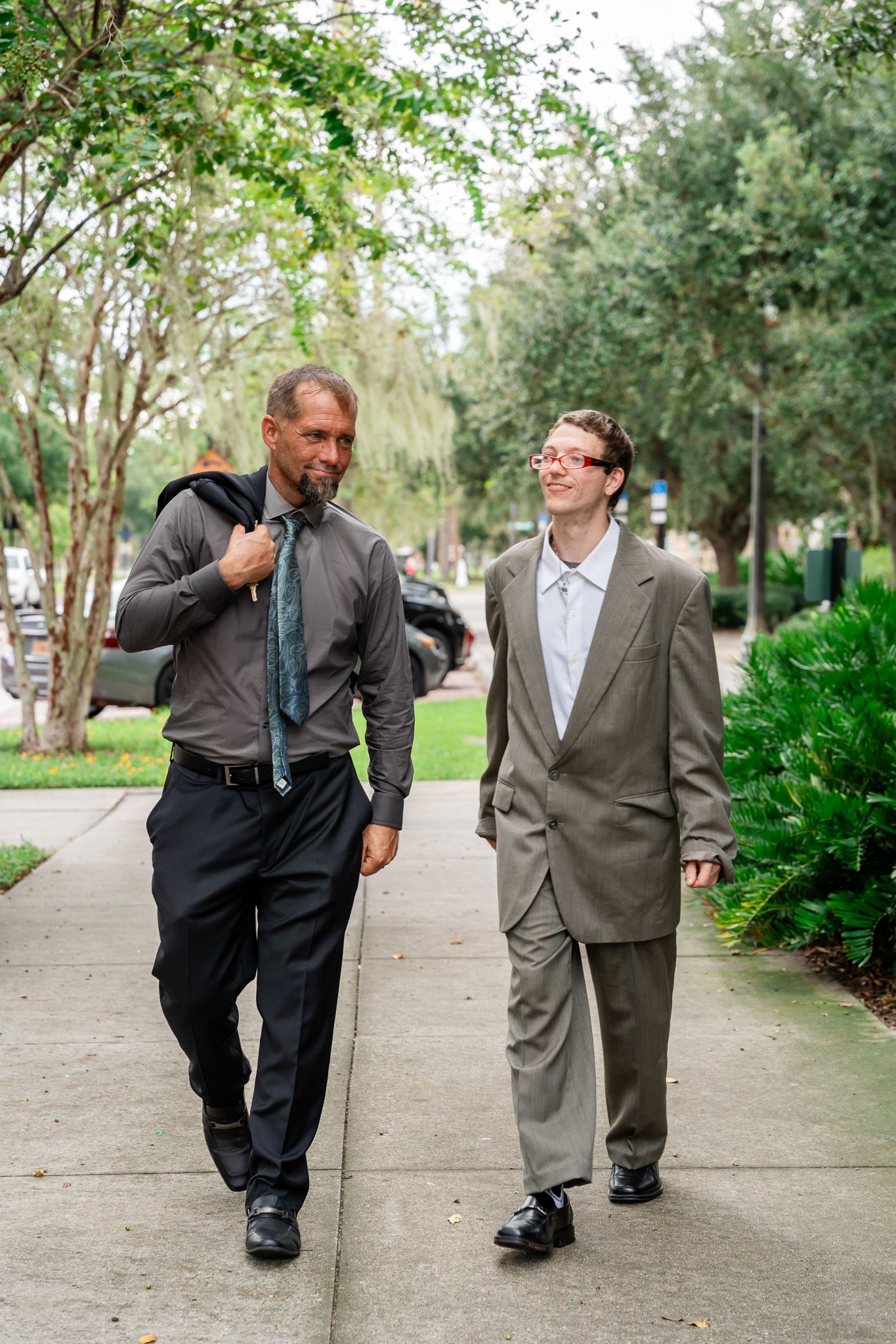 Two businessmen in suits walking together on a tree-lined sidewalk, engaged in conversation.