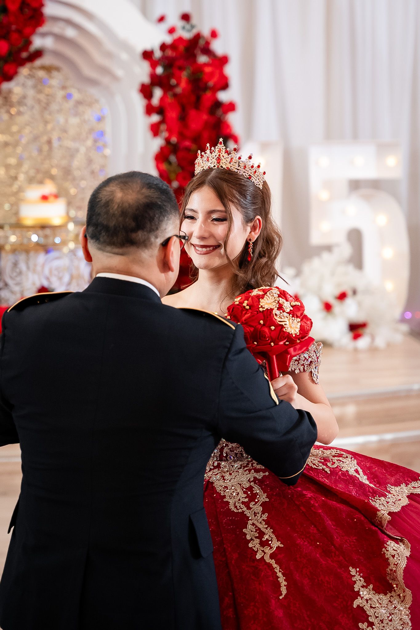 Smiling quinceañera in red gold gown and crown shares dance with father at her 15th birthday.
