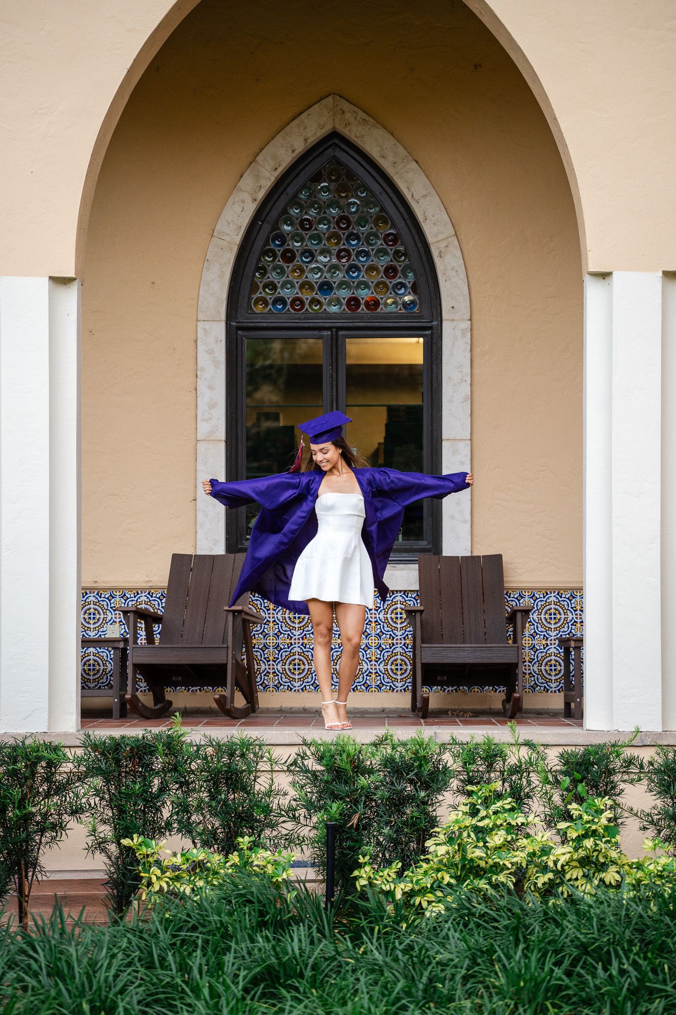A young woman in a purple graduation cap and gown over a white dress poses joyfully under an arched window.