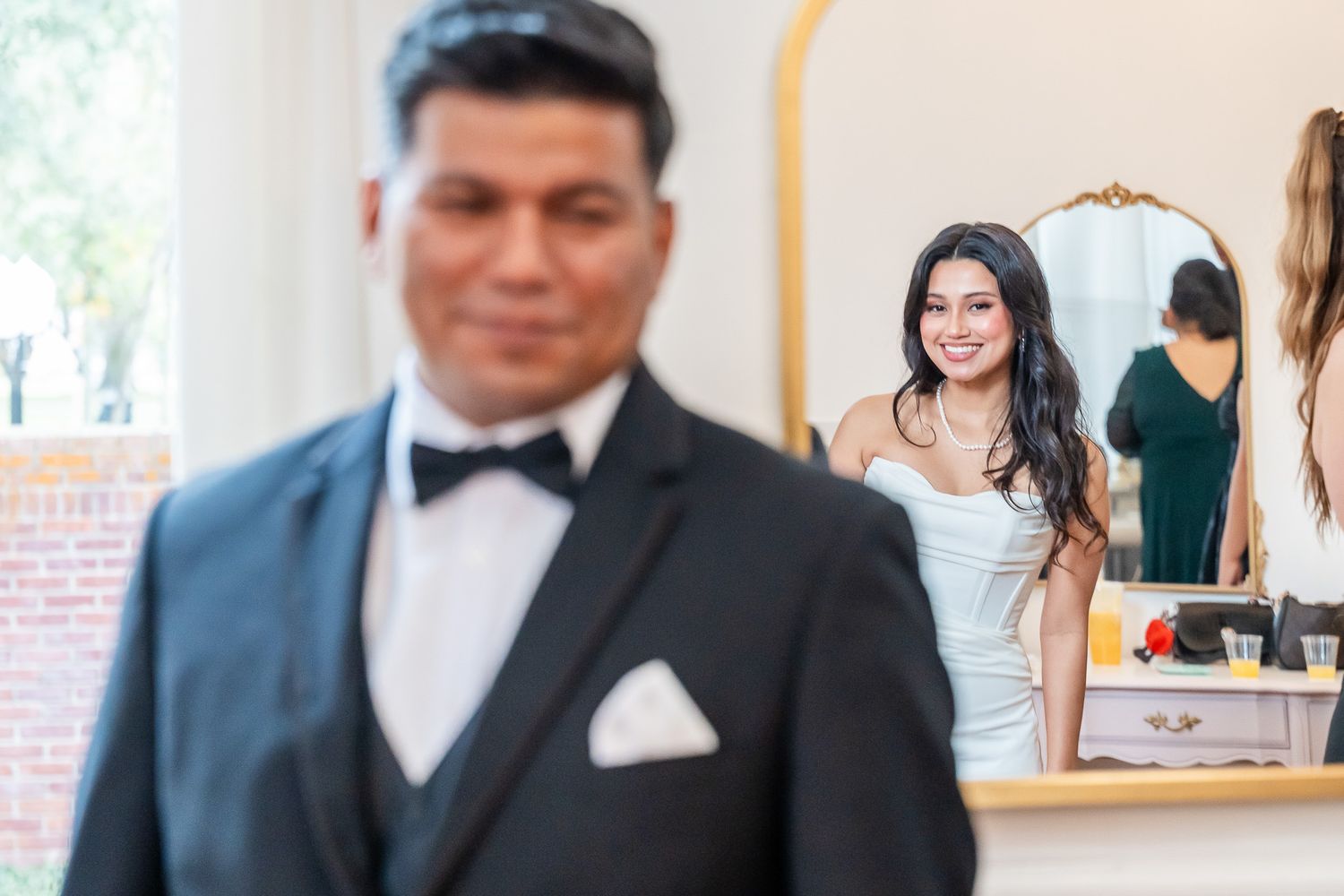 Bride smiling at groom in mirror reflection during wedding day preparations.