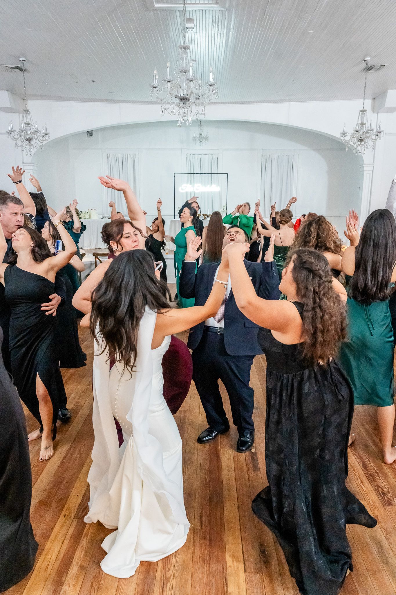 Bride and wedding guests dancing with arms raised at elegant white reception hall.