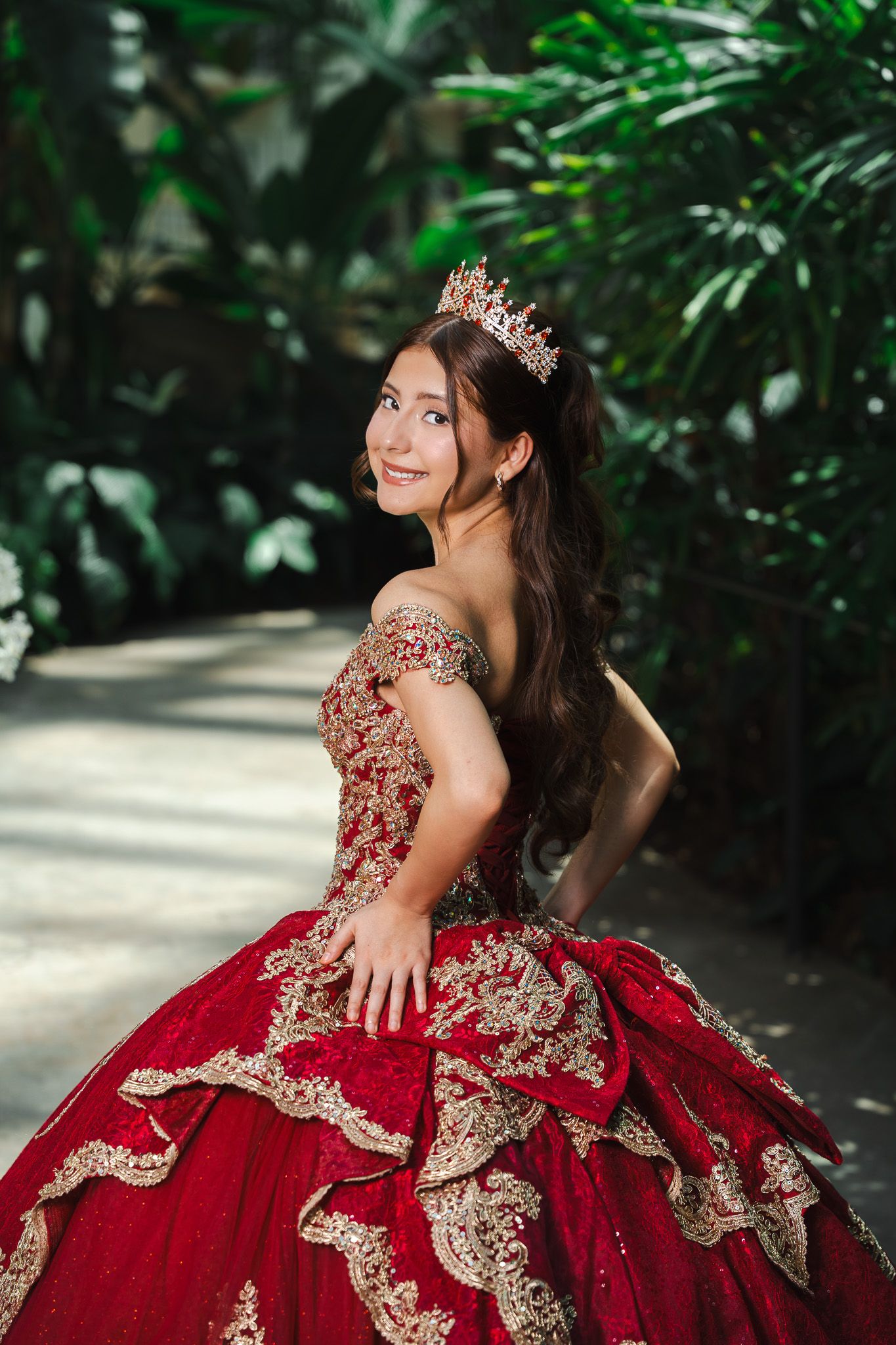 Young woman in red and gold quinceañera ball gown with crown poses gracefully in lush green garden setting.