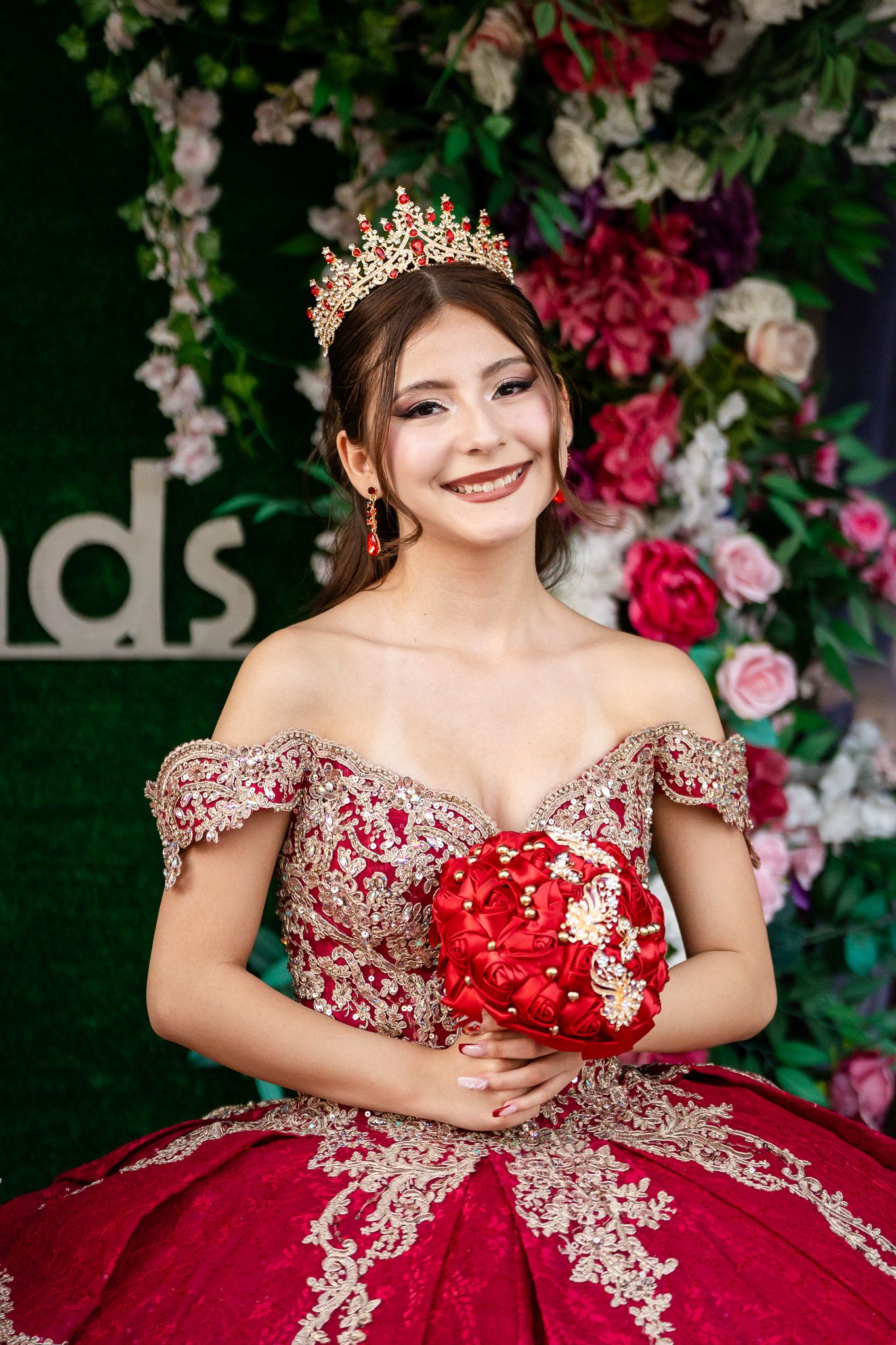 Young woman in red gold lace quinceañera gown and crown holds bouquet before floral backdrop.