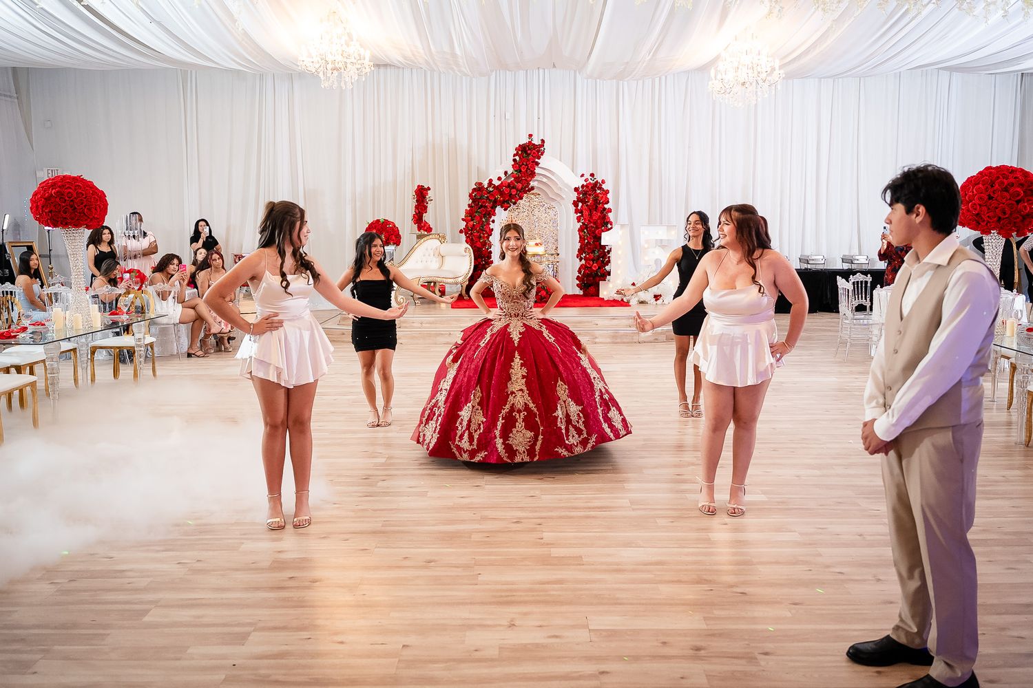 A girl in a red and gold quinceañera gown dances with her court in an elegant white decorated ballroom.