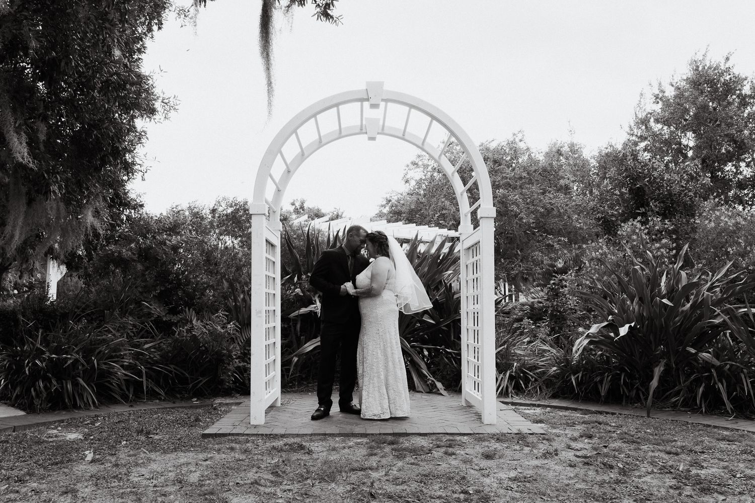 Black and white photo of a couple kissing under a white garden arch surrounded by lush greenery at their wedding ceremony.