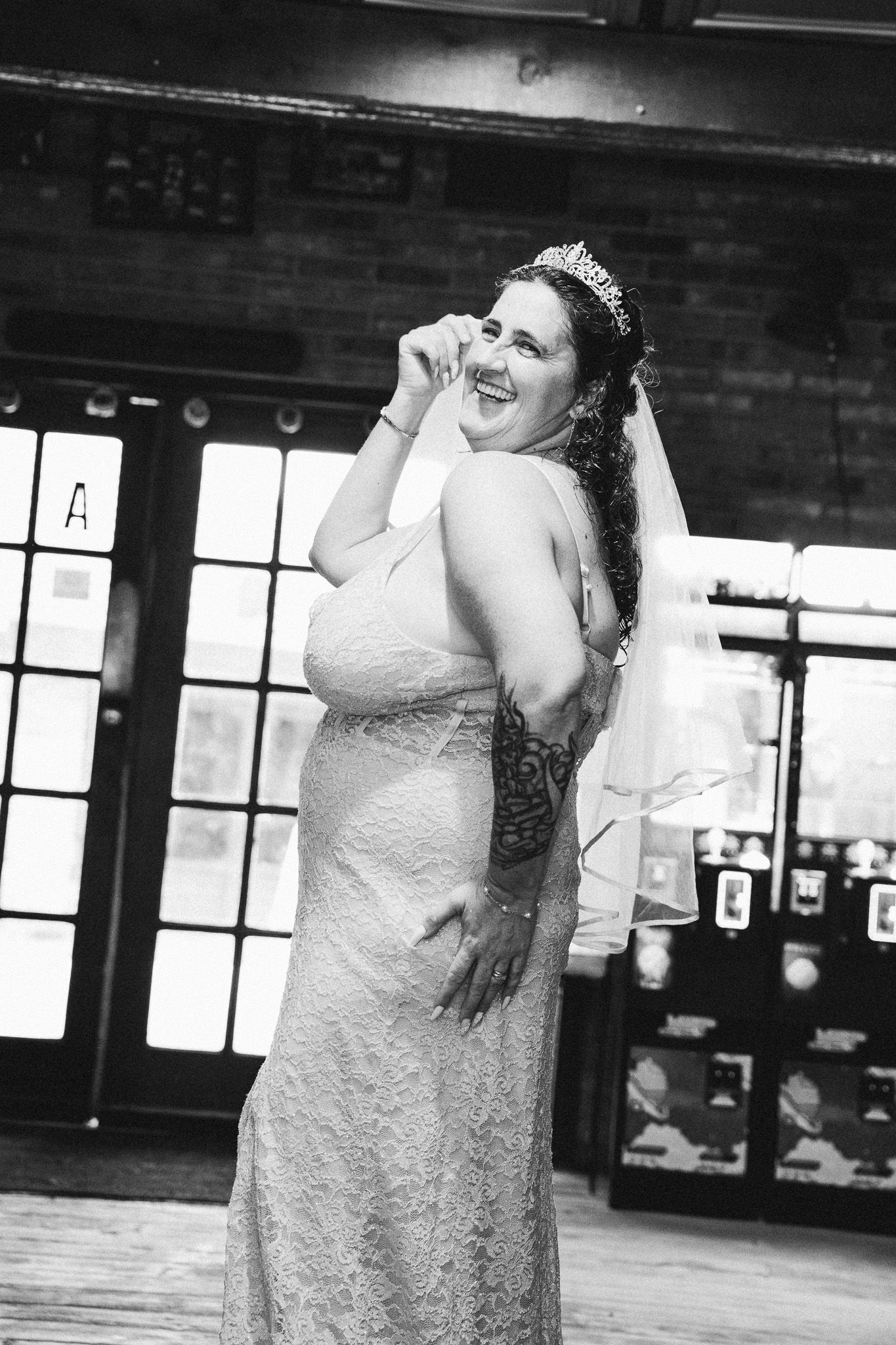 Black and white photo of a bride in lace gown and tiara, smiling joyfully inside a rustic wedding venue.