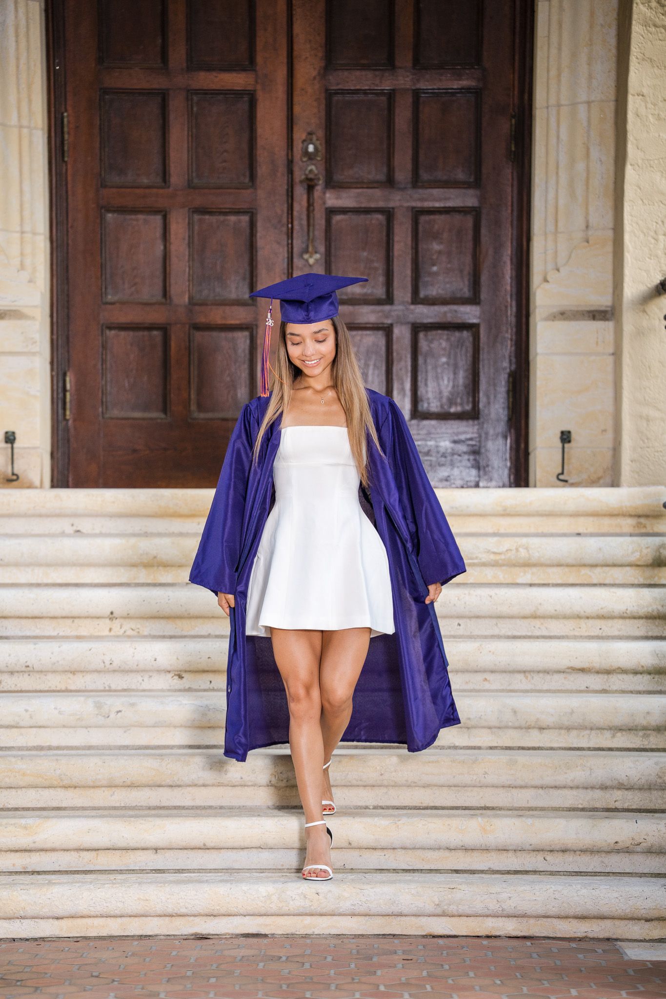 Young woman in purple graduation cap and gown over white dress walking down marble steps.