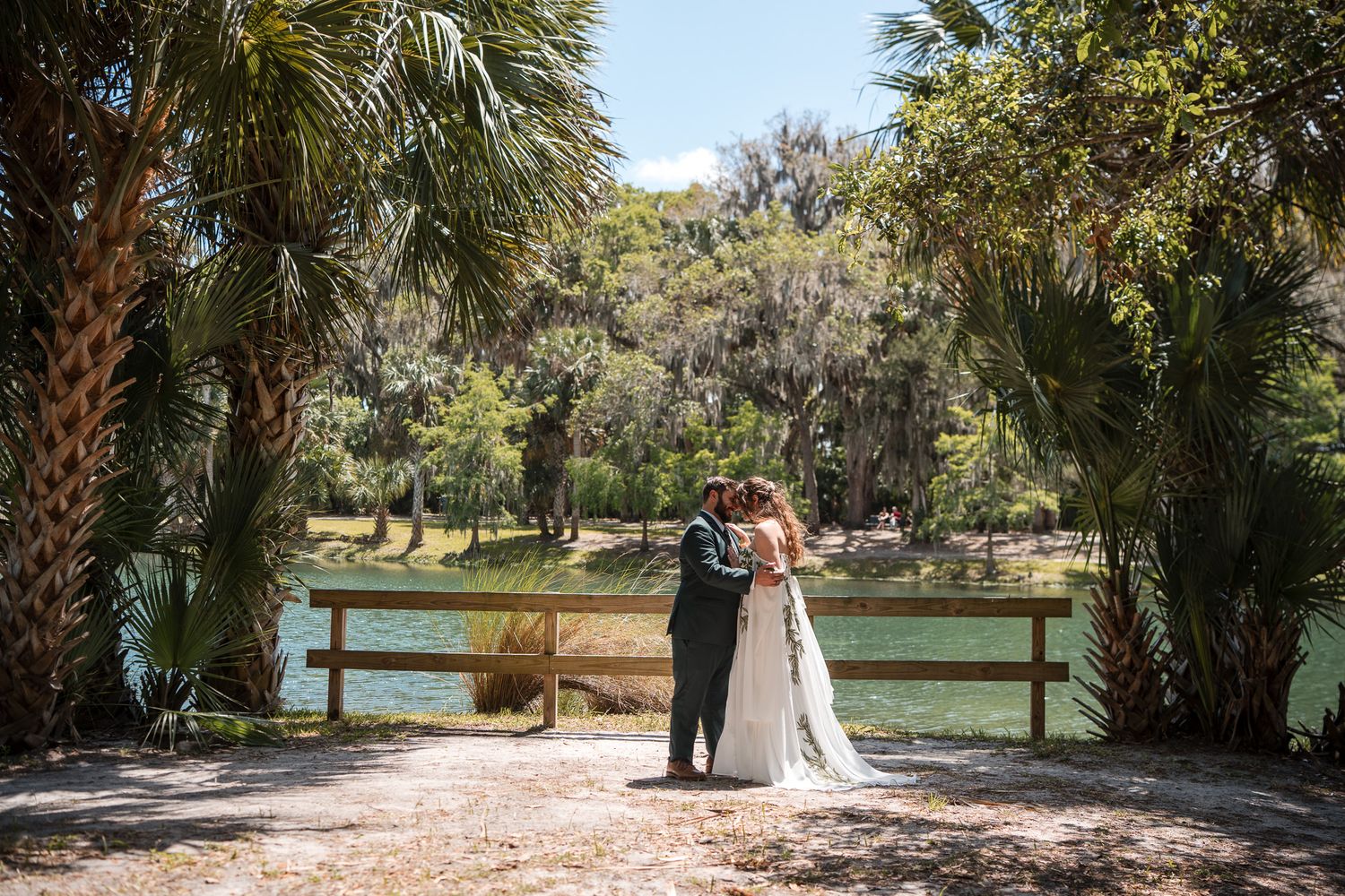 Bride and groom share a romantic kiss near a scenic lake surrounded by lush palm trees at an outdoor wedding venue.