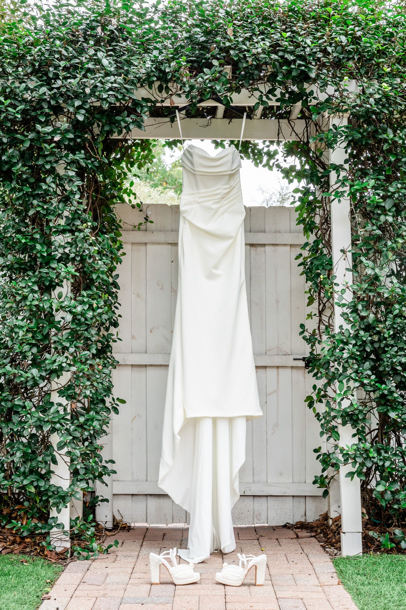 Elegant strapless white wedding gown hanging on a vine-covered garden arbor with bridal heels below.