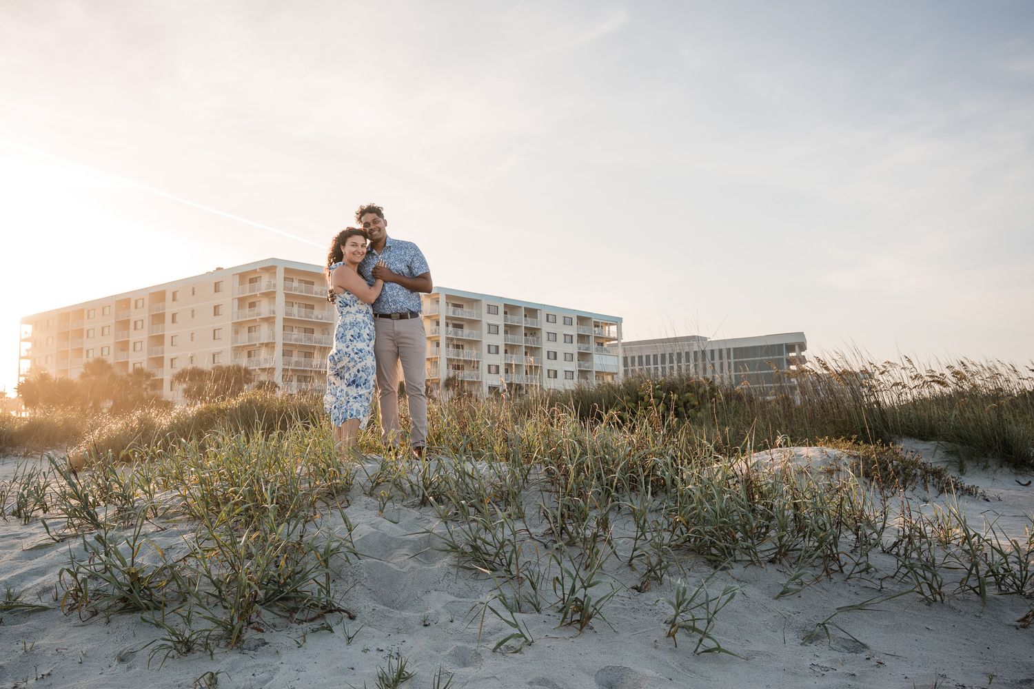 Couple embracing on sand dunes at sunset with beachfront buildings behind them on a clear evening.