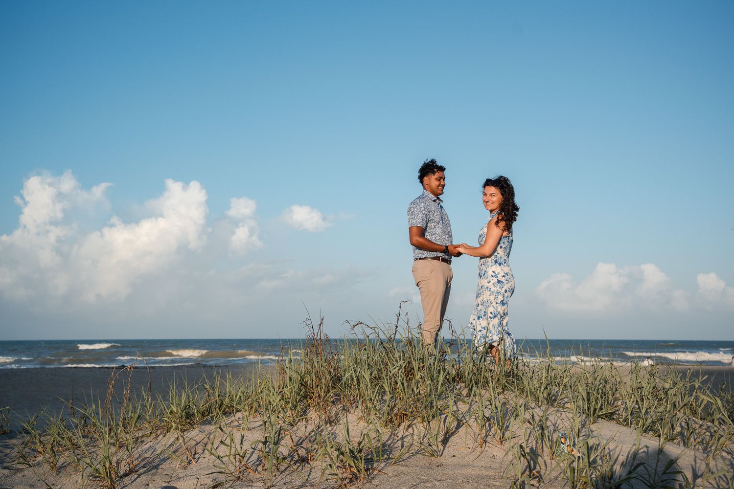 Couple holding hands on sandy beach dunes with ocean waves and blue sky in the background.