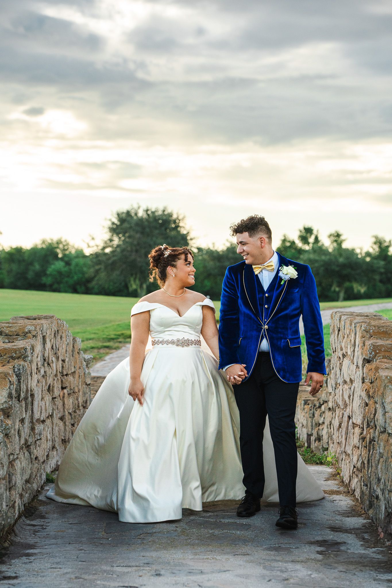 Bride in off-shoulder gown and groom in blue velvet tuxedo walk hand-in-hand on a stone bridge at sunset.
