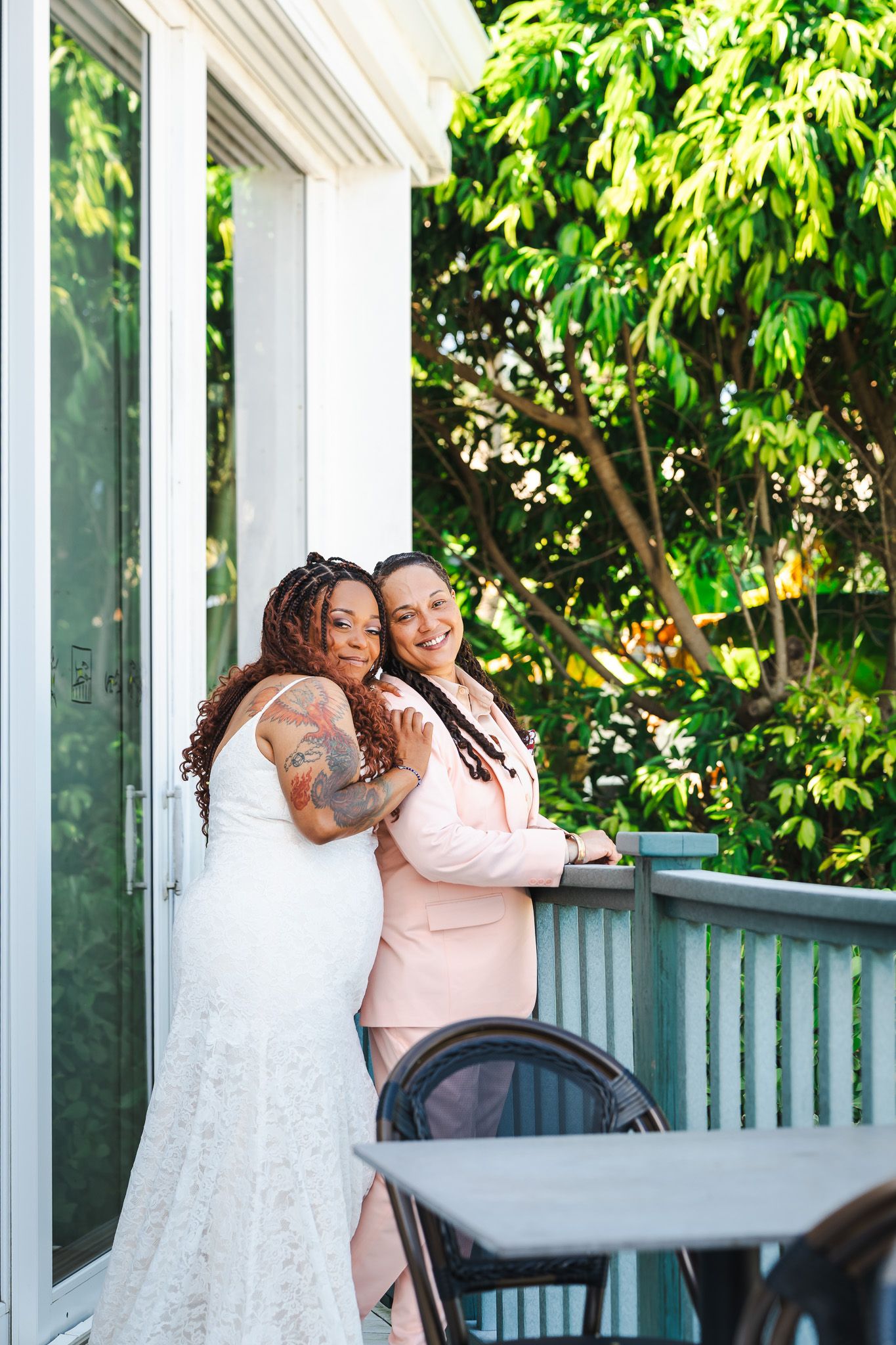 Two brides embrace on sunlit balcony surrounded by green trees in wedding attire.