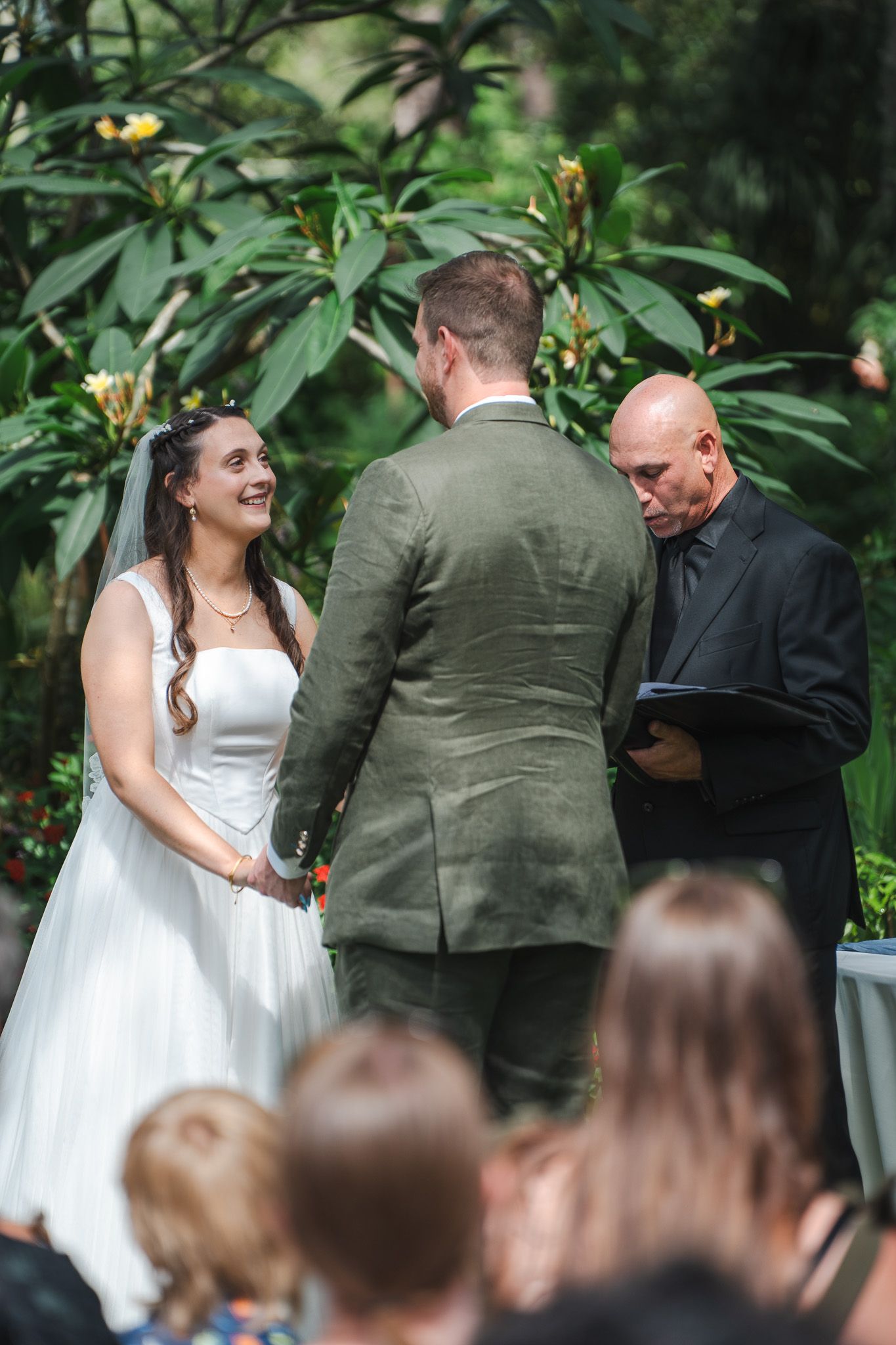 Bride laughs joyfully while exchanging vows with groom as officiant reads at garden wedding ceremony.