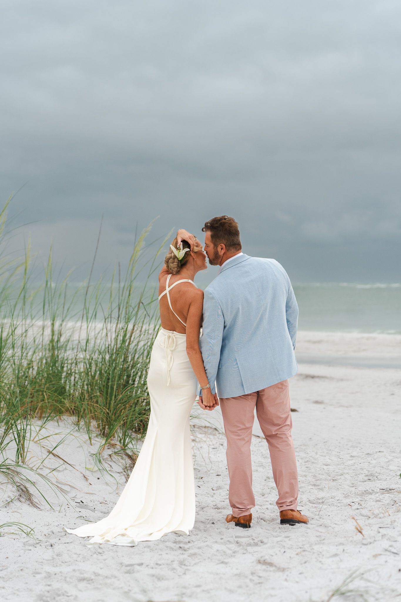 Bride and groom holding hands facing ocean with sea grass and dramatic cloudy sky at beach wedding.