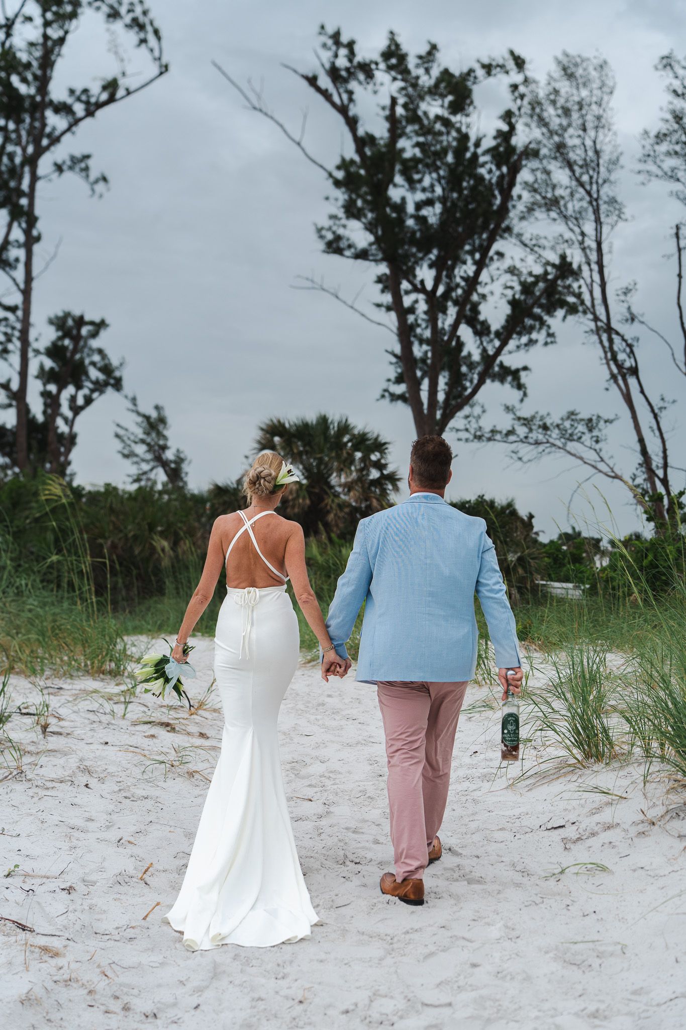 Bride and groom walking hand in hand on white sandy beach, bride wearing backless white gown.
