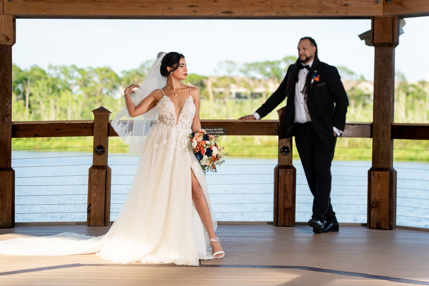 Bride in slit gown holding bouquet with groom on waterfront pavilion at sunset.