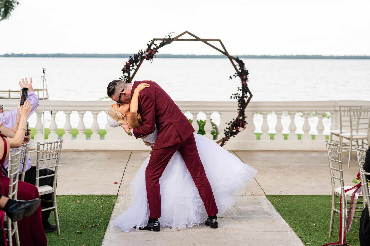 Bride and groom sharing romantic dip kiss at outdoor waterfront ceremony under circular floral arch.