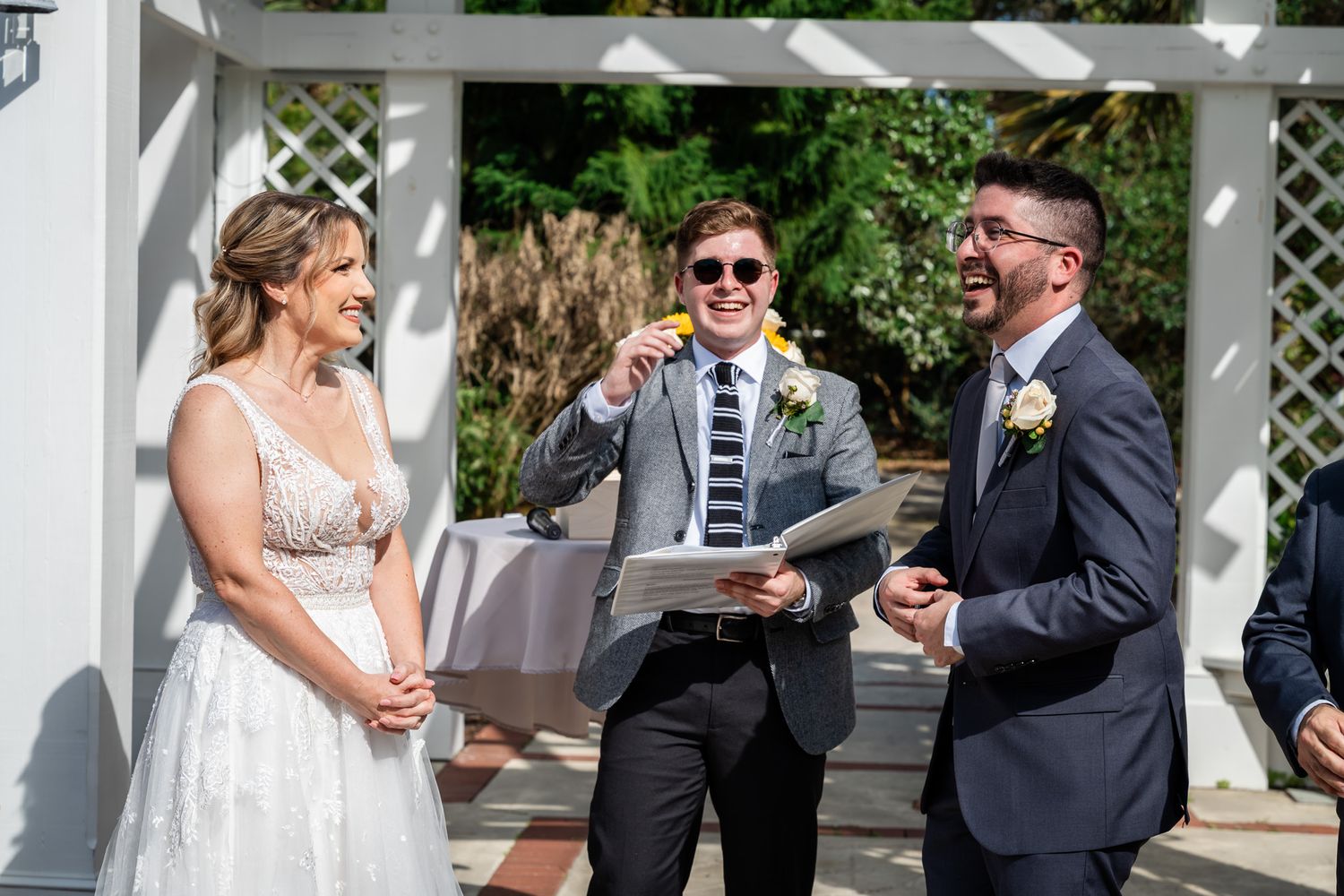 Bride and groom laugh with smiling officiant holding notes during sunny outdoor wedding ceremony at white pergola.