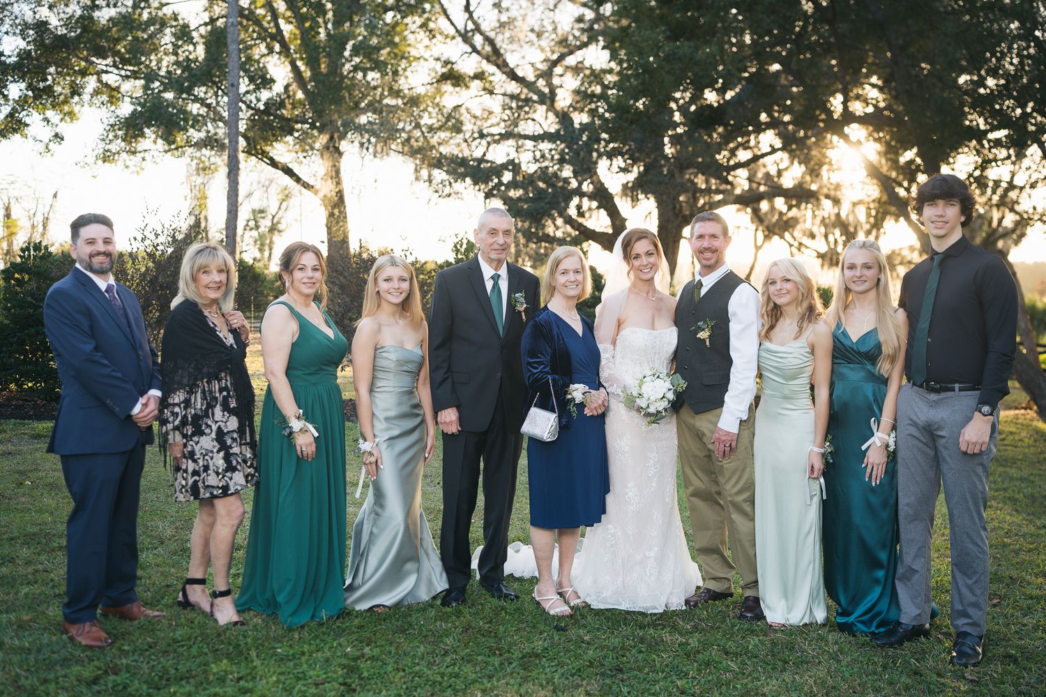 Newlyweds pose with large family group in coordinating green dresses at golden hour wedding.