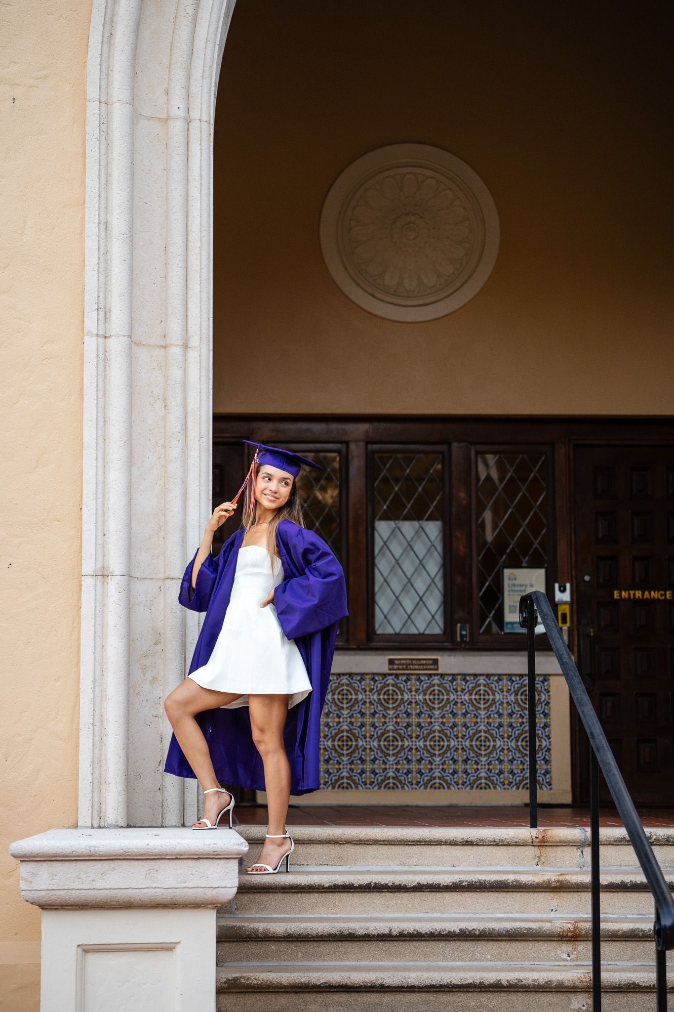 Female graduate in purple cap and gown poses elegantly on steps of a classic building entrance.