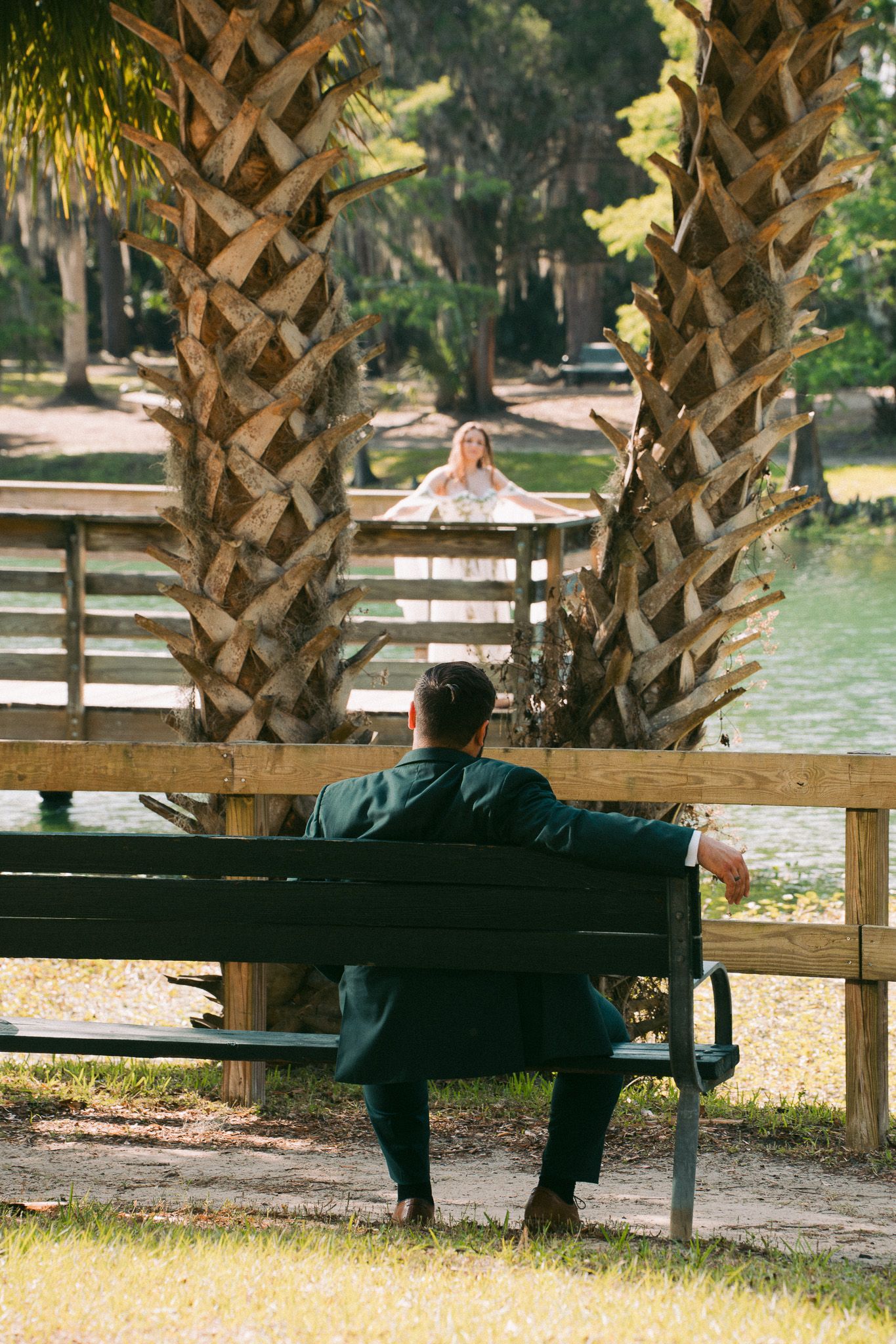 Man in green suit sits on park bench between palm trees, facing a woman standing on a wooden dock by a lake.