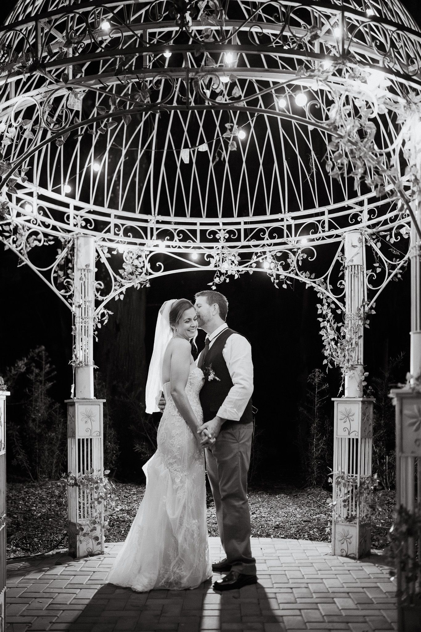 Bride and groom share a kiss under an ornate white gazebo decorated with string lights at night.