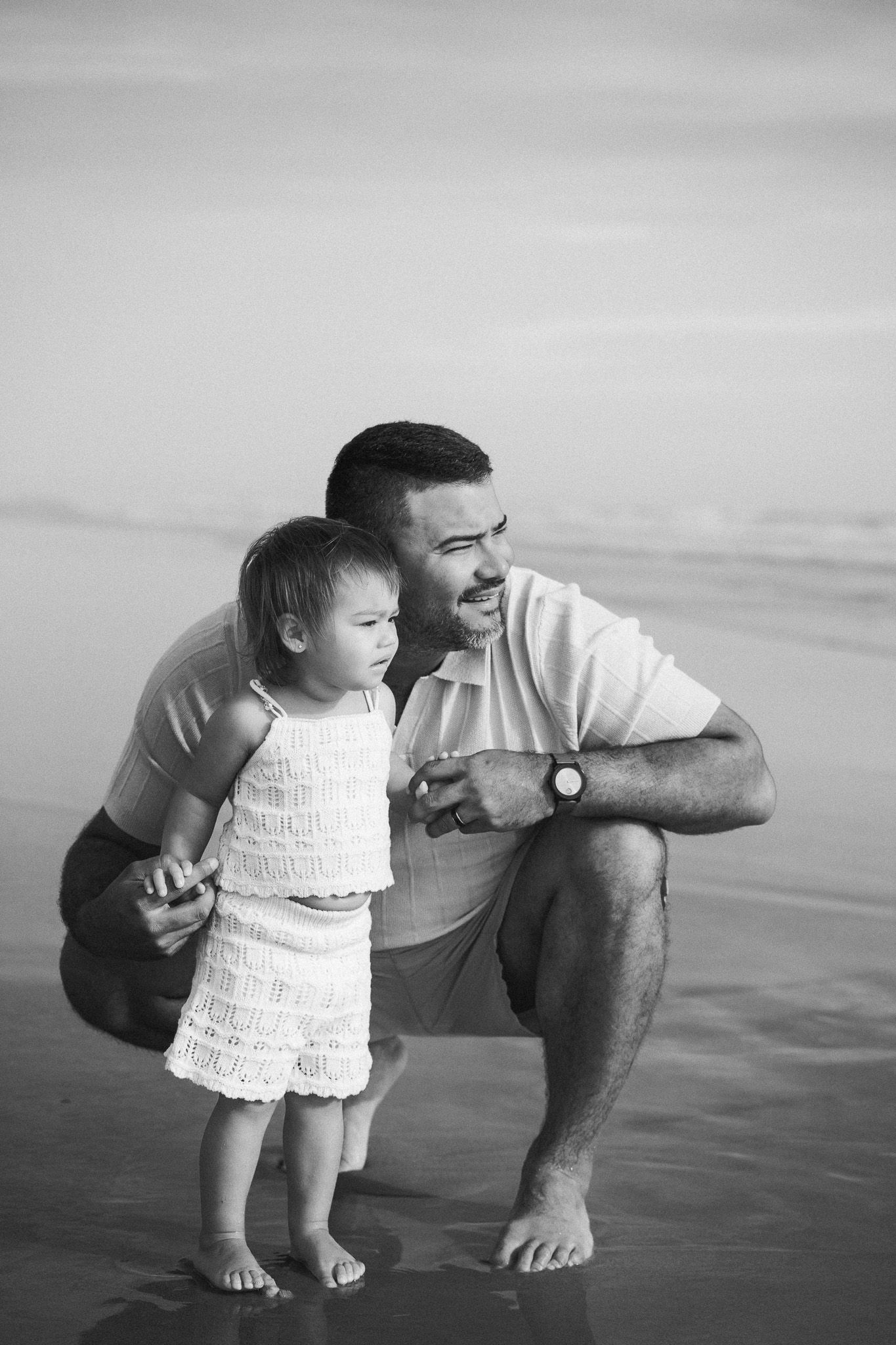Father crouching beside toddler daughter on beach shore in black and white photo.