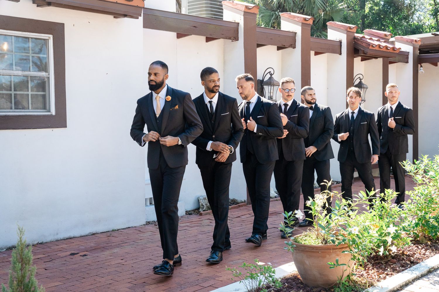 Groom leads groomsmen in black suits walking along a brick pathway beside a white stucco wall.