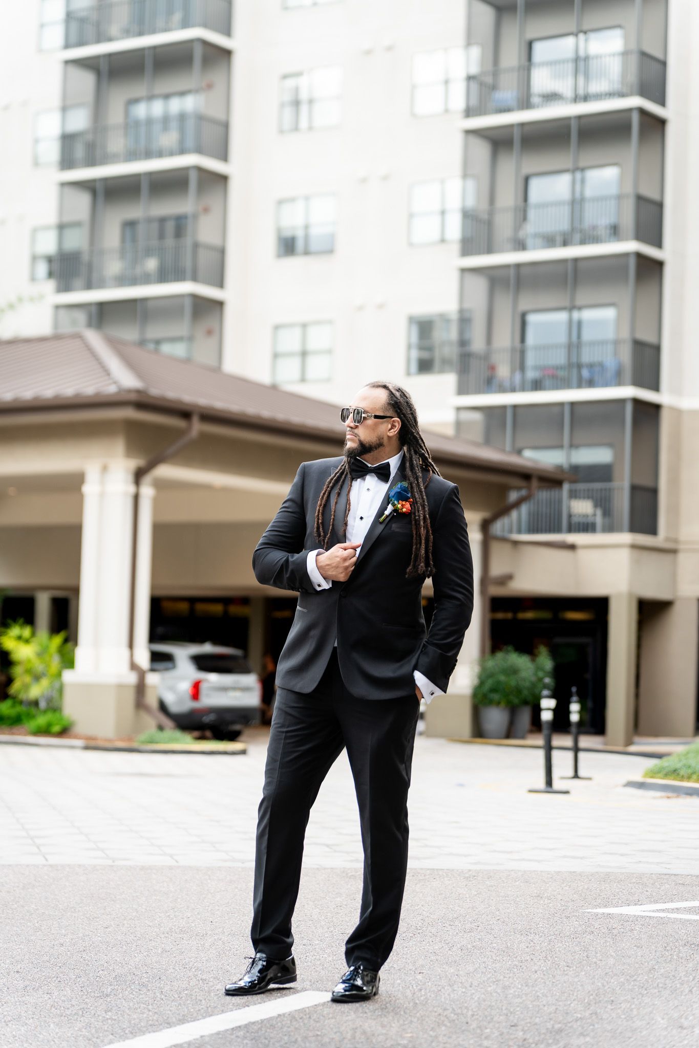 Stylish groom in black tuxedo with dreadlocks and sunglasses poses outside a hotel building.