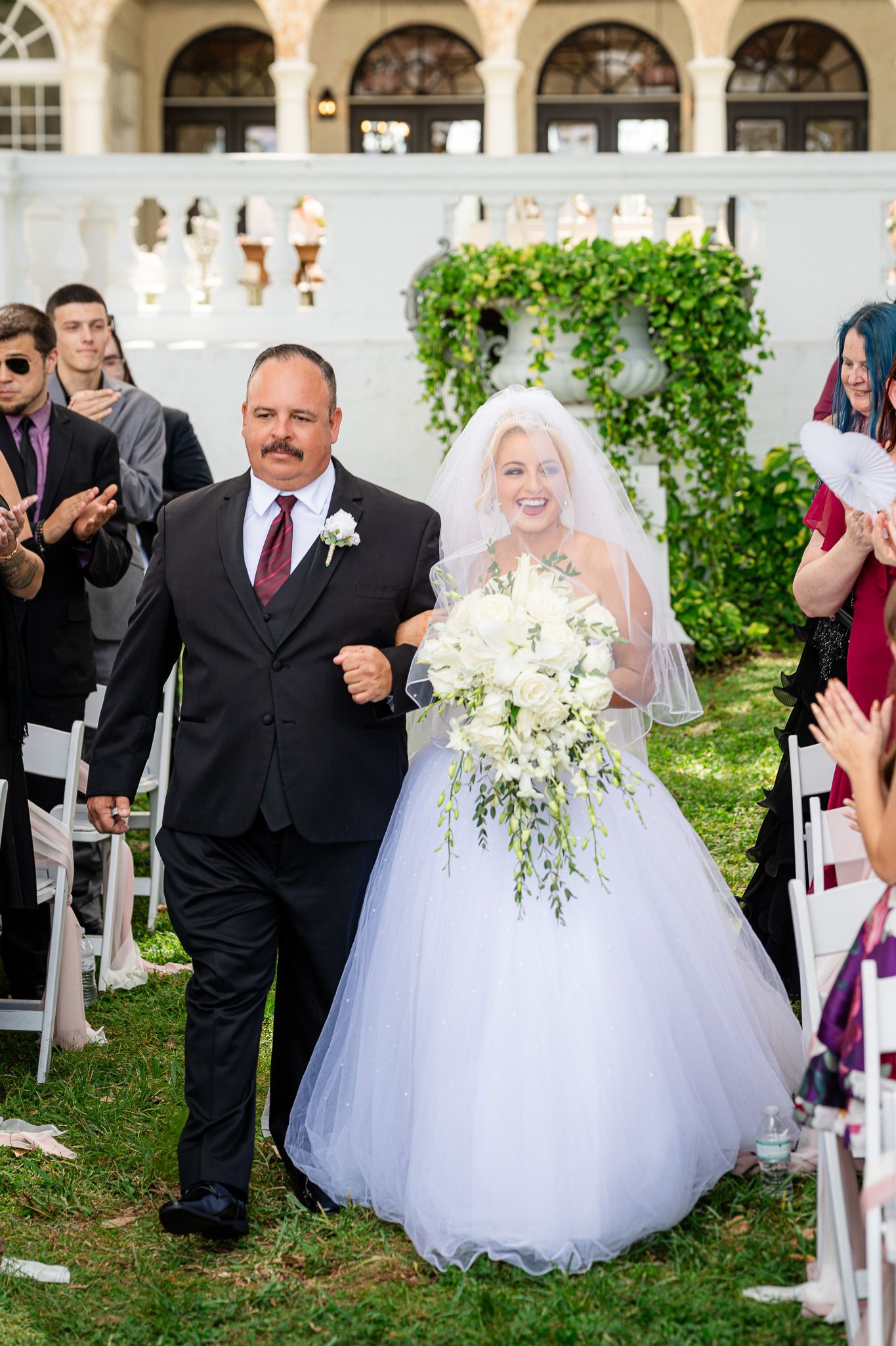 Smiling bride holding white cascading bouquet walks down aisle with father at outdoor ceremony.