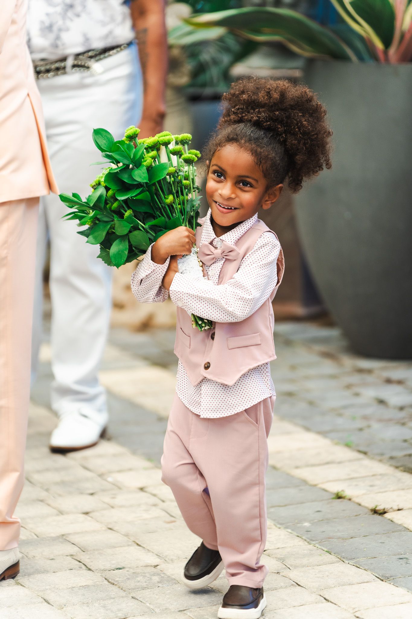 Smiling young ring bearer in pink suit holding a bouquet of green flowers at an outdoor wedding.