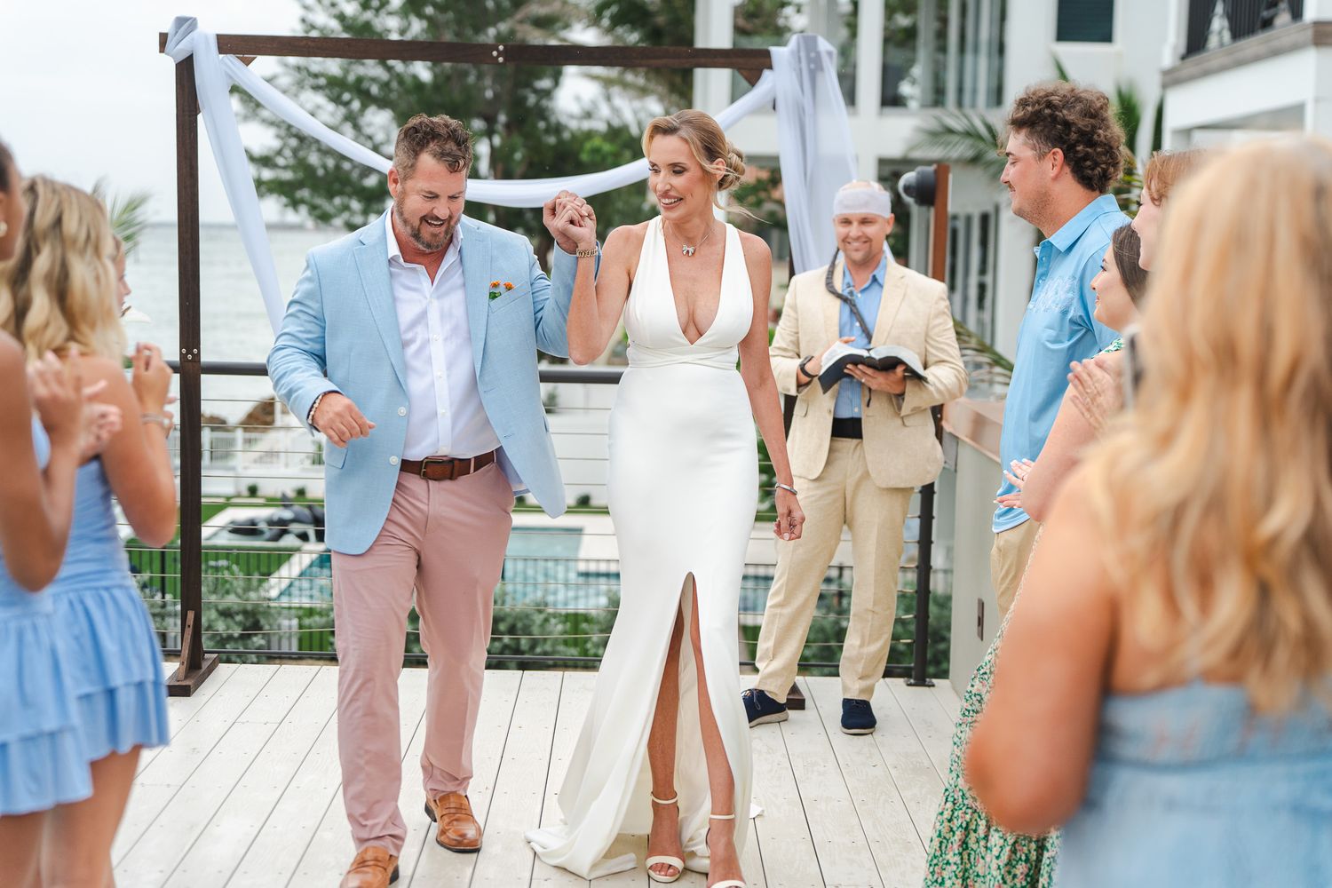 Newlyweds walk joyfully hand in hand past cheering guests after outdoor beachside wedding ceremony.