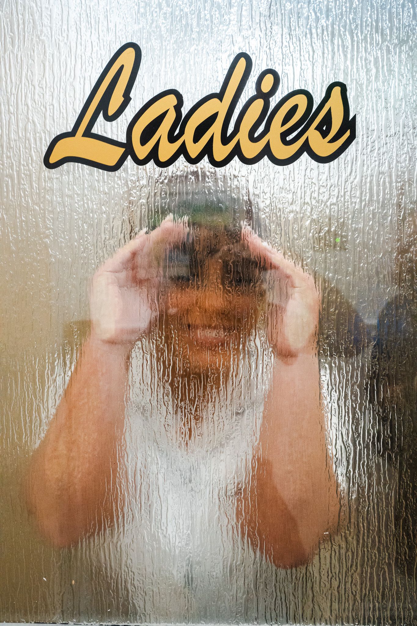 Smiling bride in white dress peeking through frosted glass Ladies restroom door.