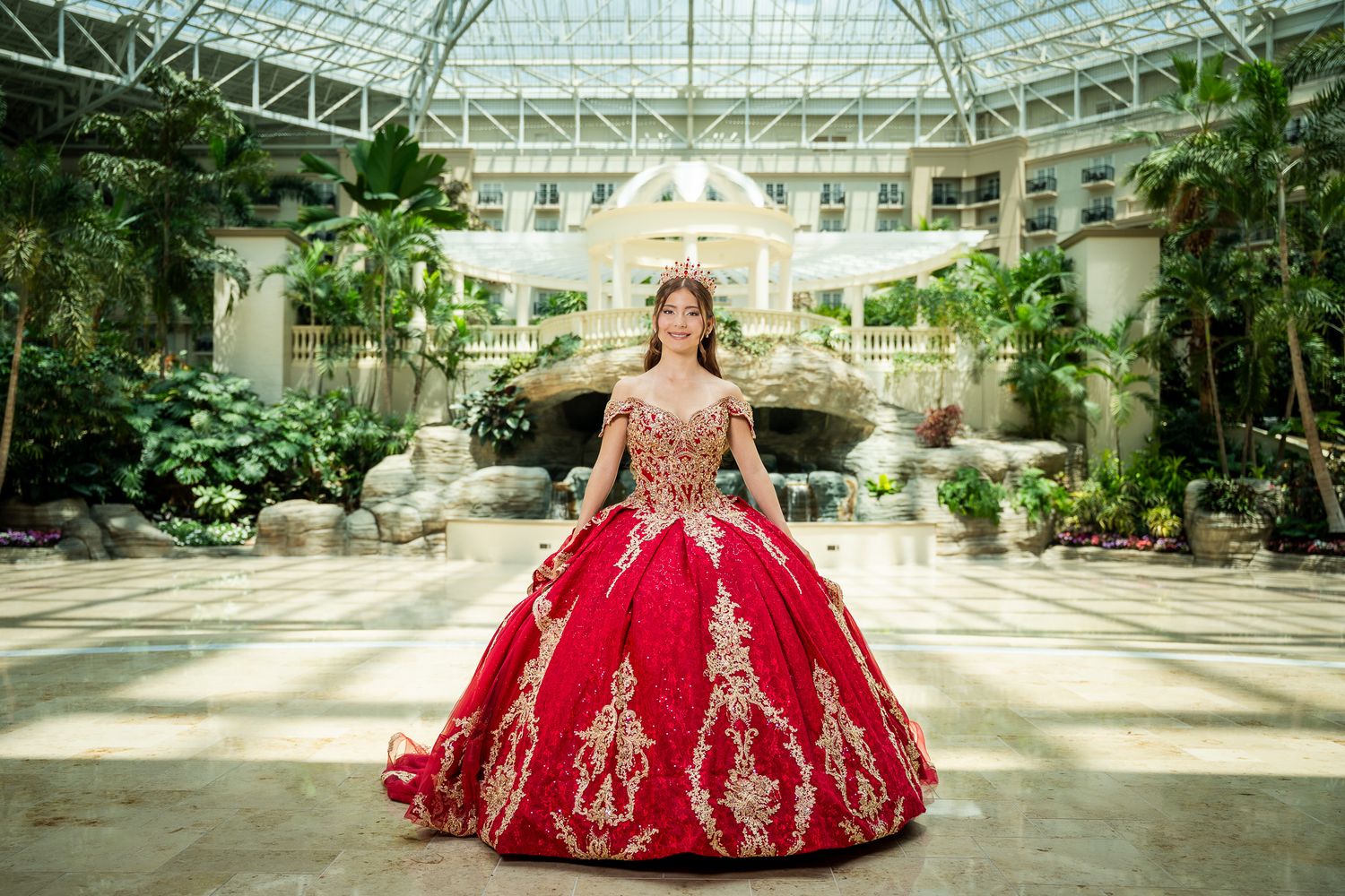 Young woman in a red and gold quinceañera ball gown poses in an elegant indoor garden atrium.