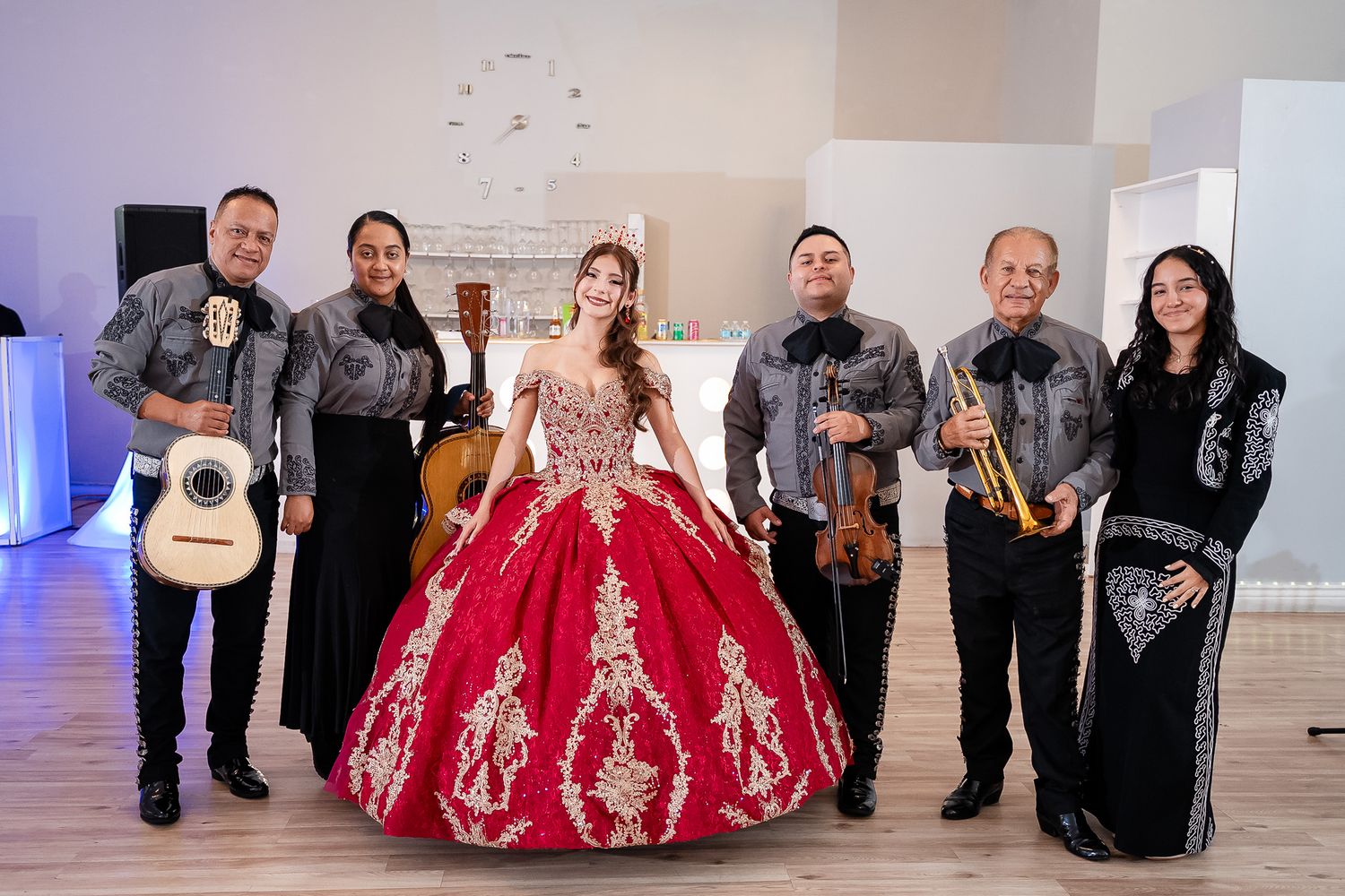 A quinceañera girl in a red gown poses with a mariachi band holding guitars and violin in a decorated venue.