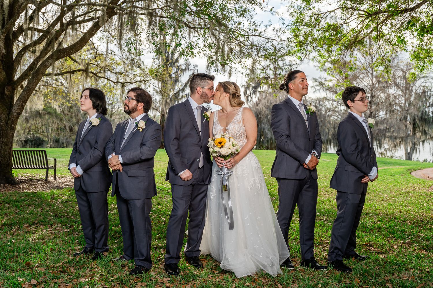Wedding party with bride and groom kissing flanked by four groomsmen in gray suits under moss-covered oak trees.