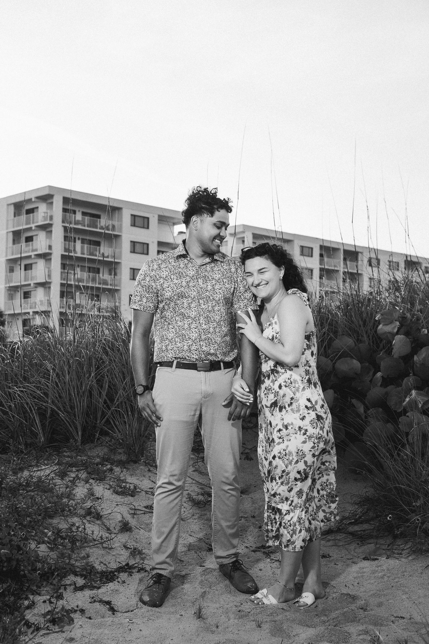Black and white photo of a couple holding hands and smiling on a sandy beach with dune grass and a hotel behind them.