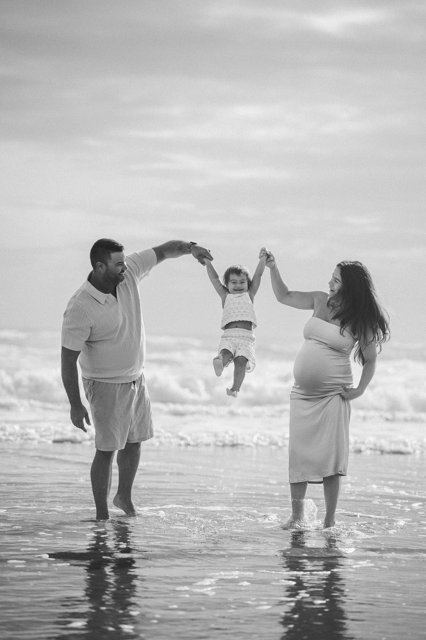 Black and white photo of couple swinging toddler between them on beach with ocean waves.