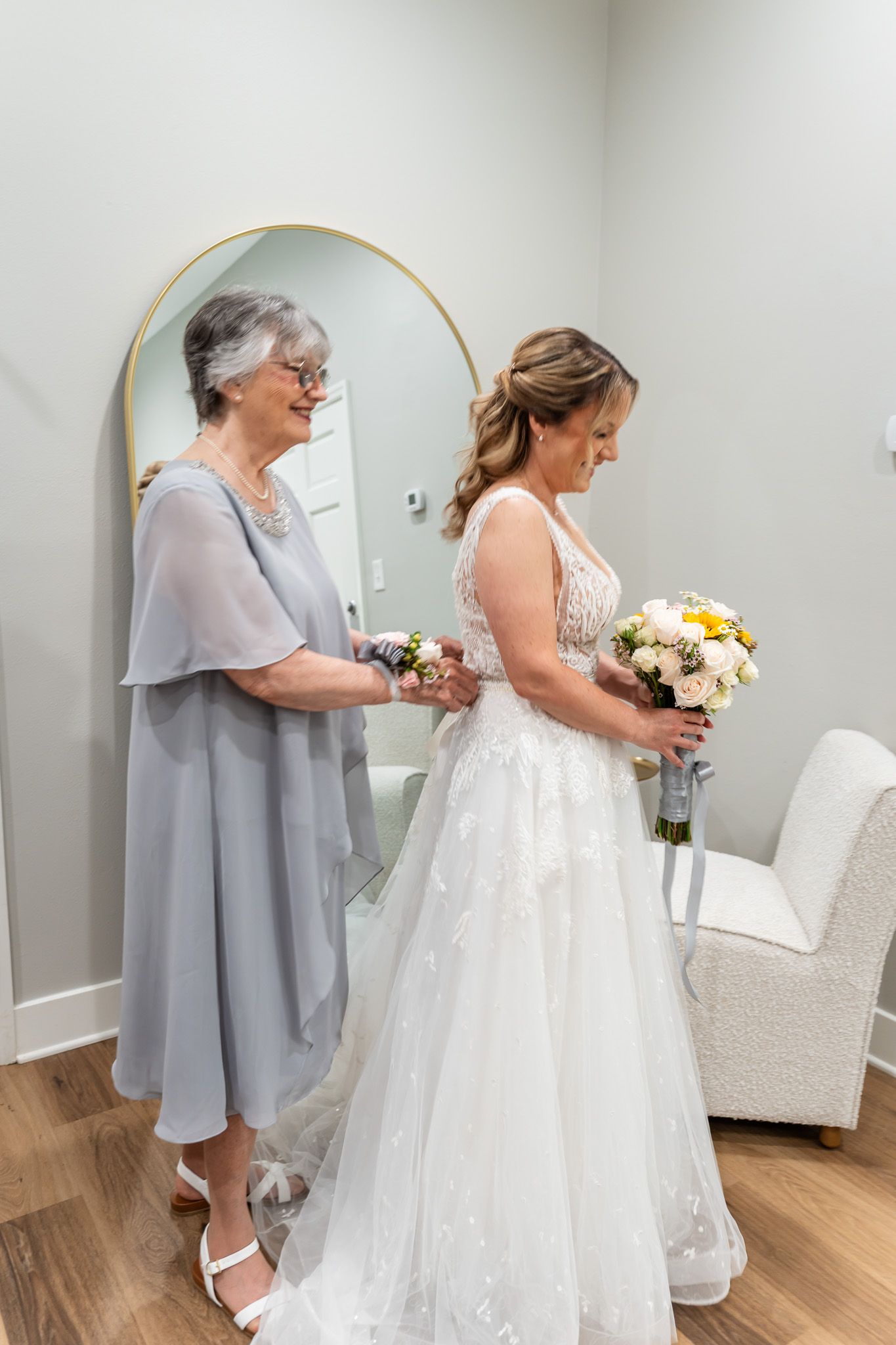 Mother helps bride with dress details as she holds a yellow and white floral bouquet.
