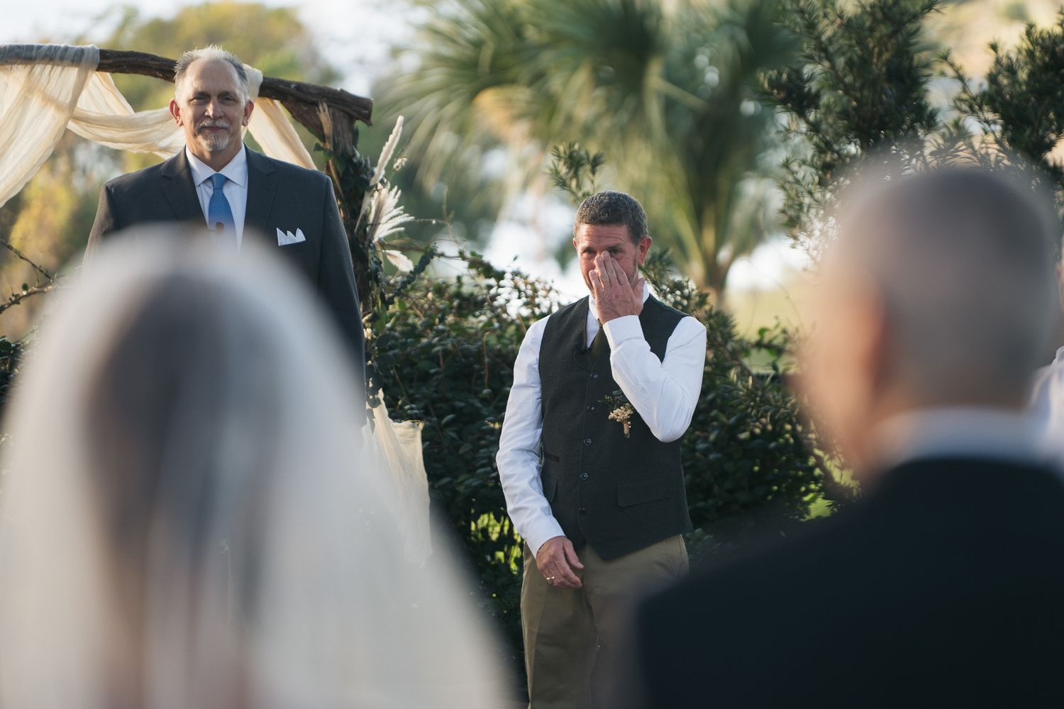 Emotional groom wiping tears as bride walks down the aisle at outdoor wedding ceremony.